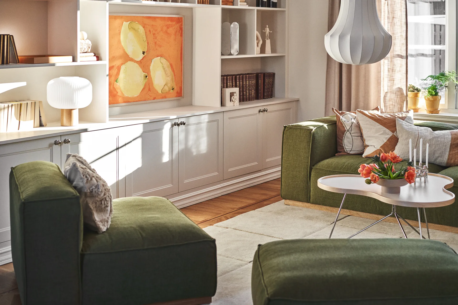 Soggiorno soleggiato con divani verdi, un tavolino bianco con tulipani arancioni e candele, e mensole bianche con opere d'arte.