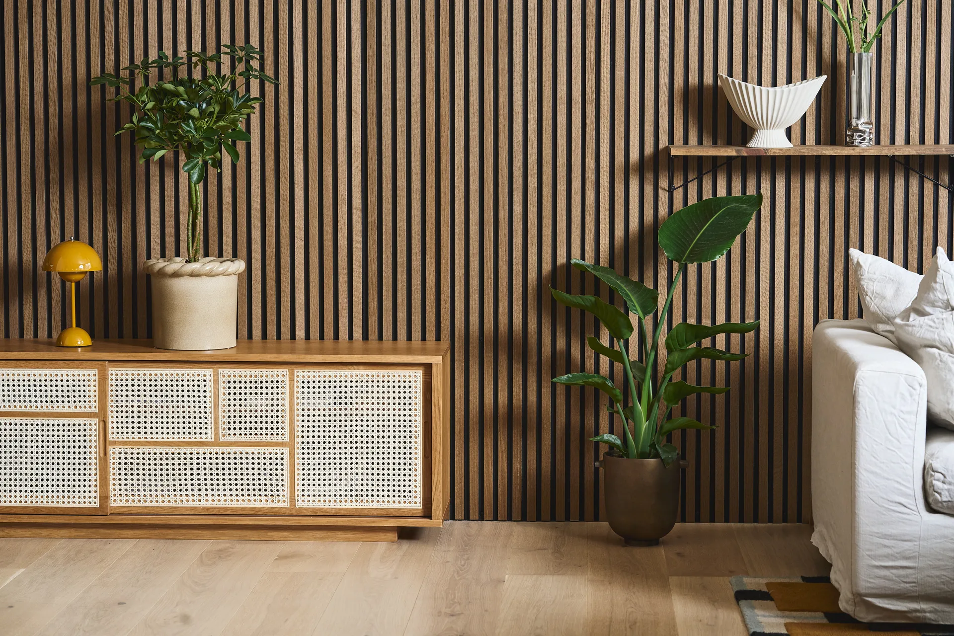 Soggiorno moderno con parete a doghe in legno, mobile in rattan, lampada gialla, piante e divano bianco.