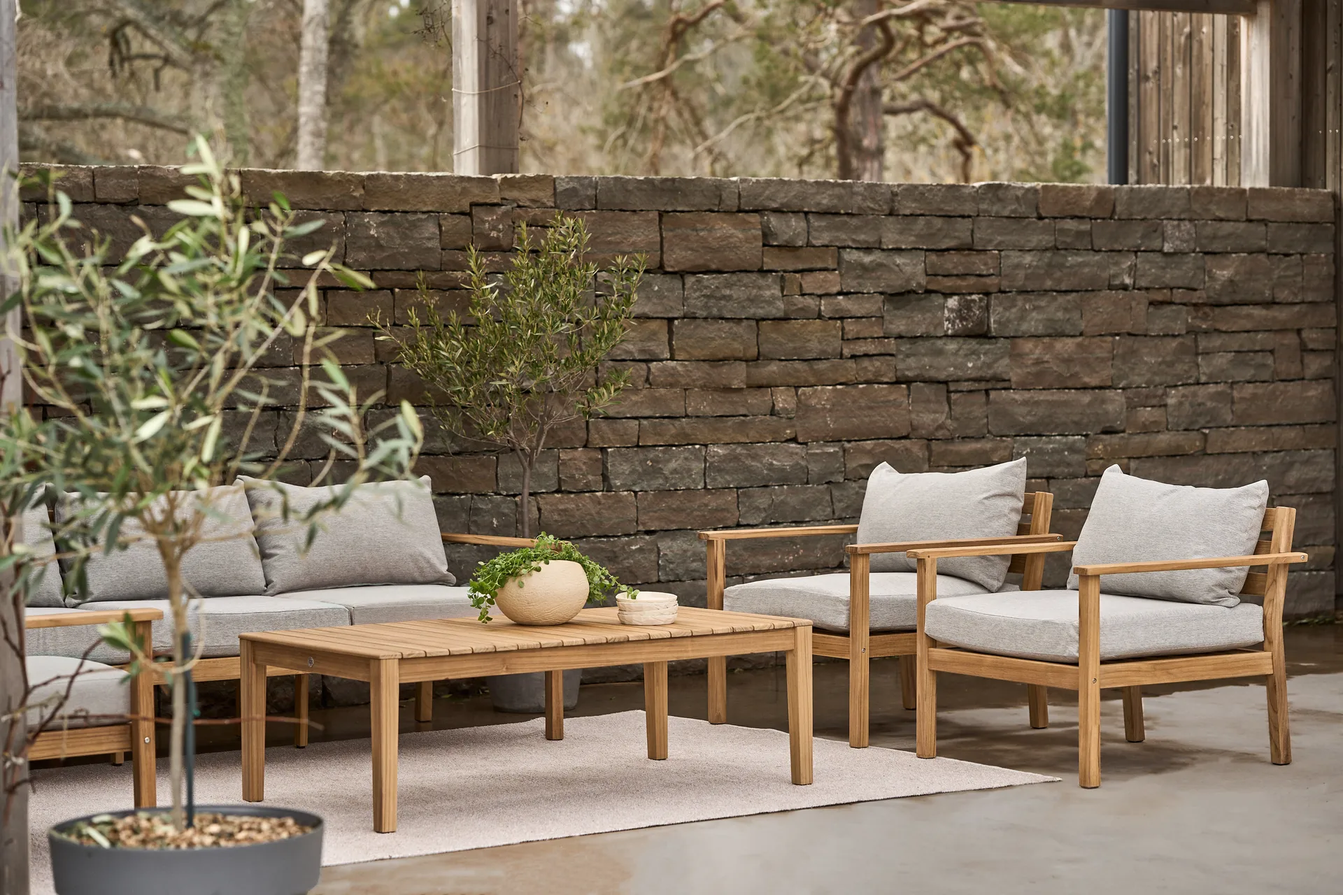 Set da giardino in legno con cuscini grigi e tavolino basso davanti a un muro di pietra.