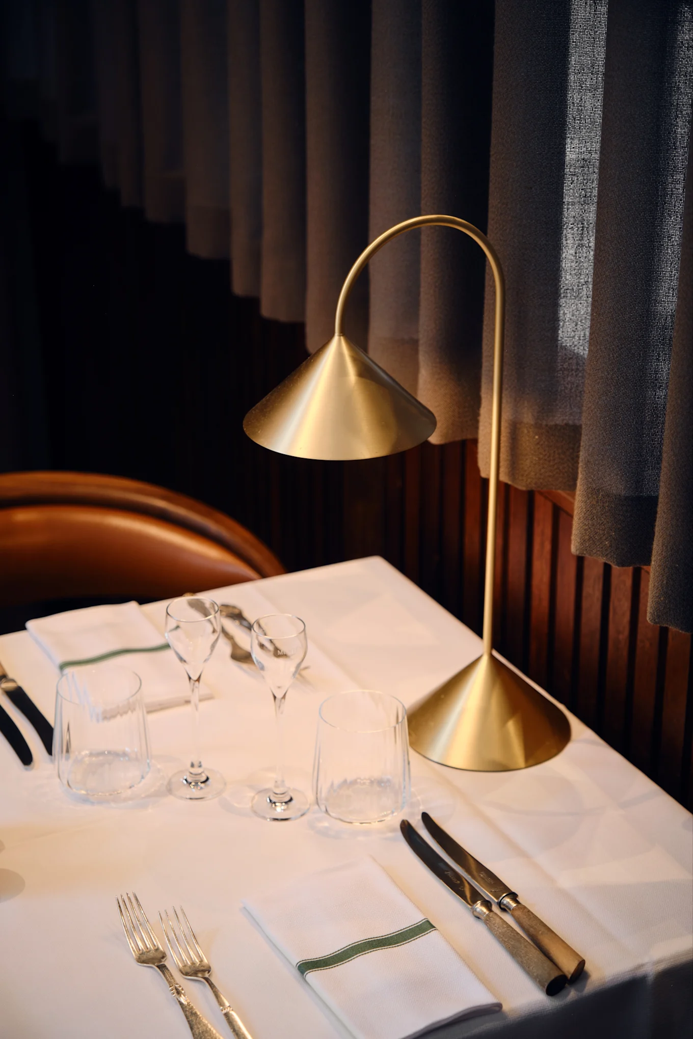 Una moderna lampada da tavolo dorata illumina un tavolo da ristorante apparecchiato con biancheria bianca, bicchieri, posate e un tovagliolo a striscia verde.