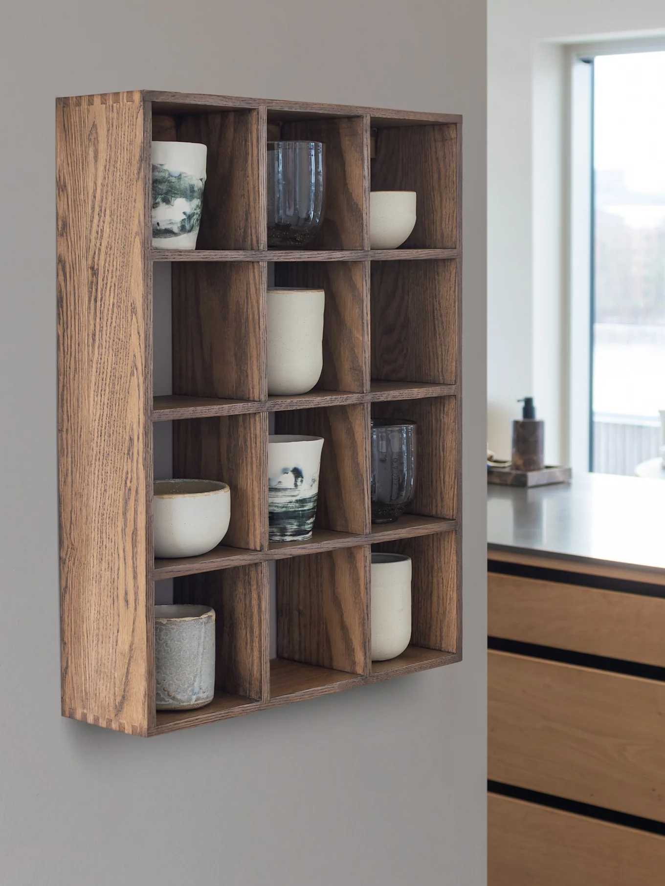 Scaffale a muro in legno con 12 scomparti contenenti tazze e mug in ceramica. Una cucina moderna e una finestra sono sullo sfondo.
