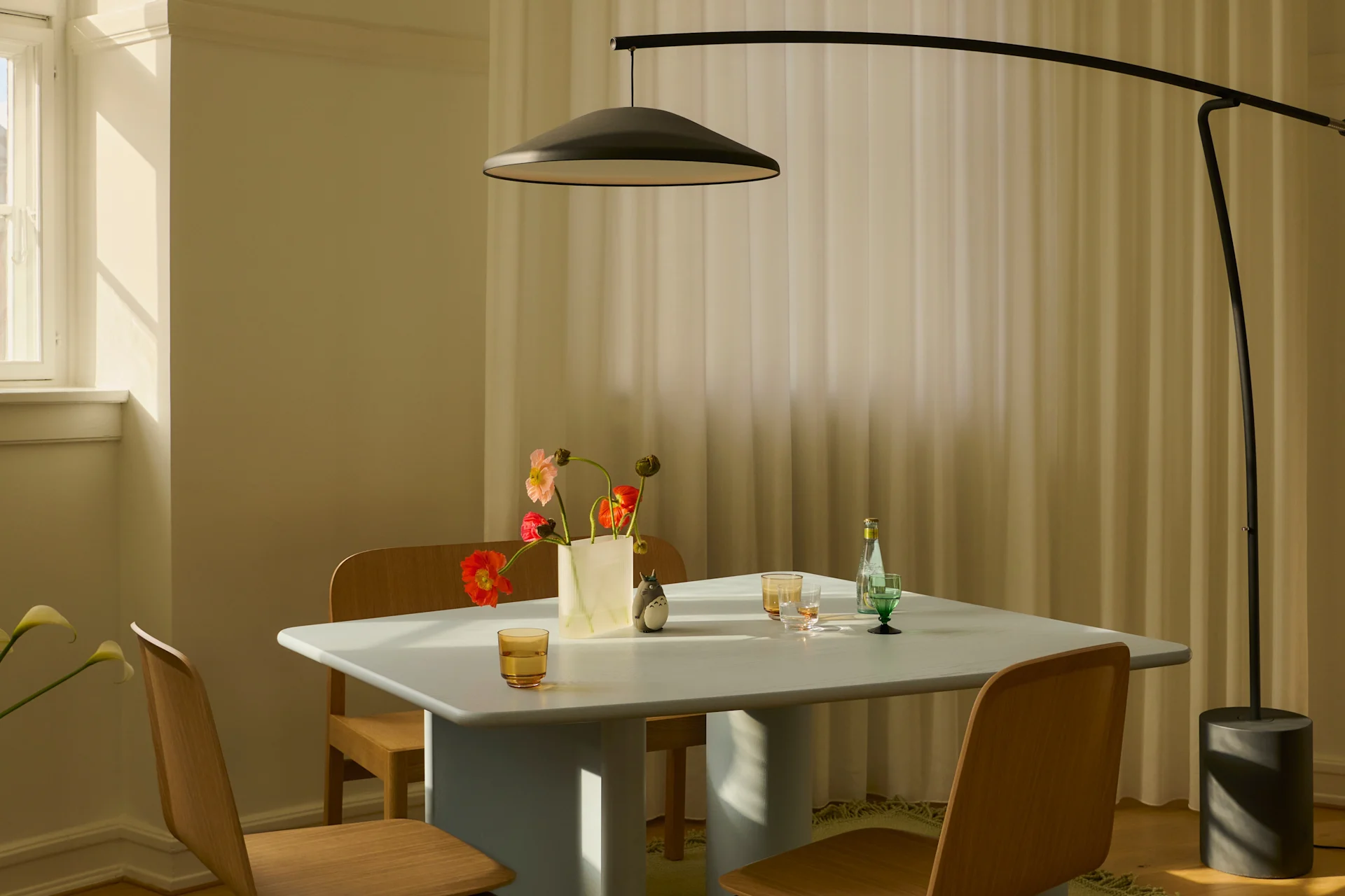 Sala da pranzo moderna con tavolo azzurro, sedie in legno, lampada ad arco nera e vaso di fiori colorati al sole.
