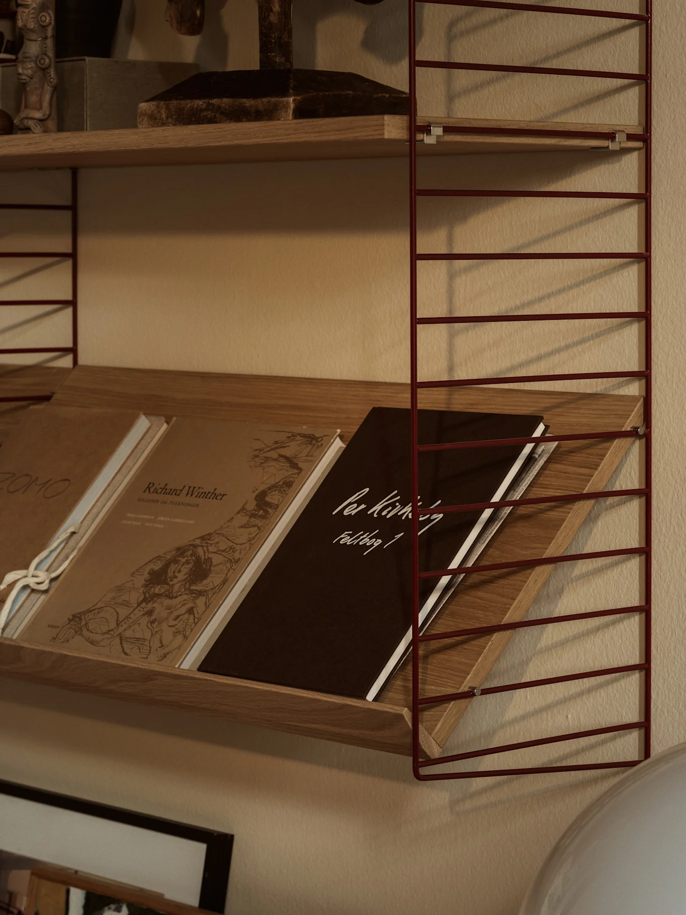 Una mensola a muro con struttura in filo metallico rosso e ripiani in legno. Il ripiano inferiore contiene libri inclinati, tra cui uno marrone scuro con 'Per Kirkeby Feldbog 1'.