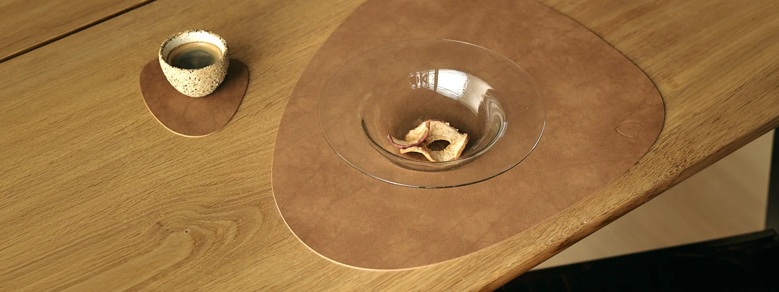 Tazza da caffè su piccolo tappetino marrone, ciotola con mele essiccate su grande tappetino marrone, su tavolo di legno.