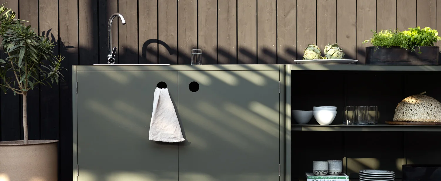 Cucina esterna con mobili verde salvia, lavello, erbe aromatiche e una pianta in vaso contro una parete di legno scuro.