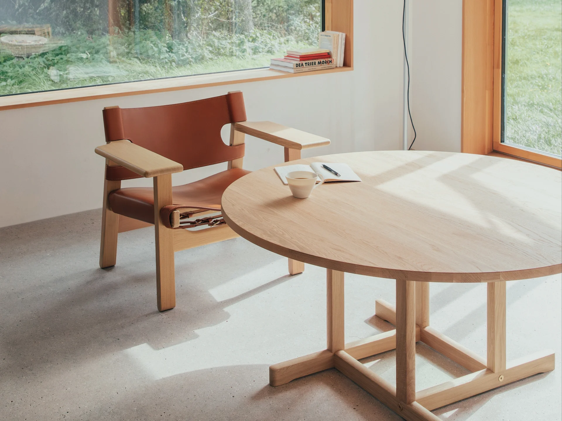 Stanza luminosa con poltrona in pelle marrone, tavolo rotondo in legno chiaro con tazza e taccuino, illuminata da finestra con vista sulla vegetazione.