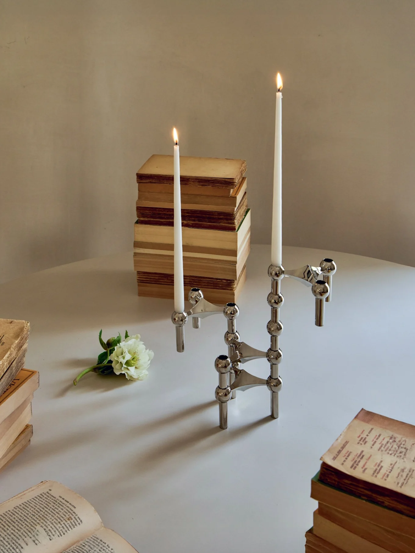Un portacandele modulare argentato con due candele accese, libri e un fiore bianco su un tavolo.
