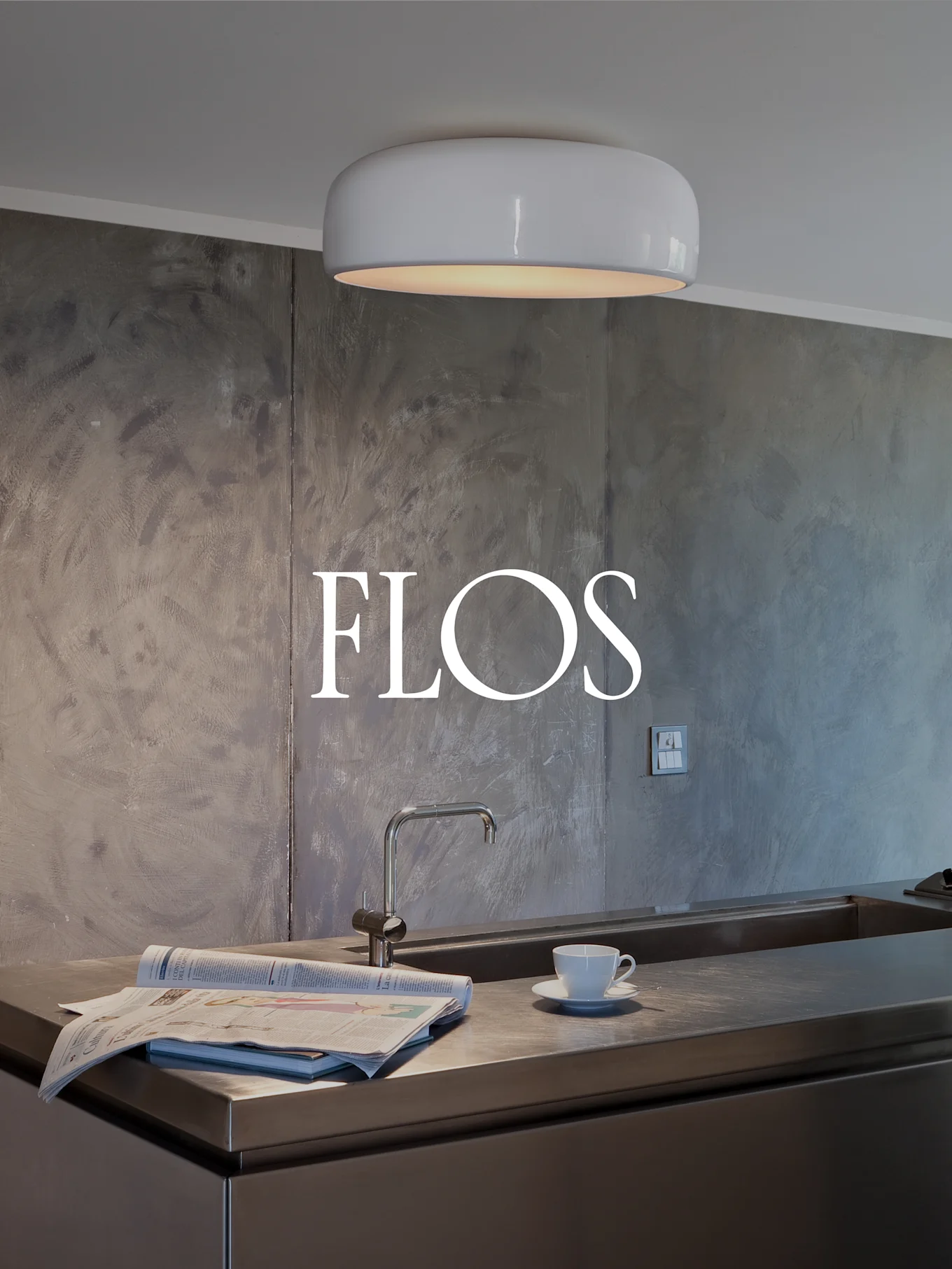 Cucina moderna con plafoniera bianca e arrotondata, bancone in acciaio inossidabile con lavello, giornale e tazza da tè. Logo FLOS sulla parete testurizzata.