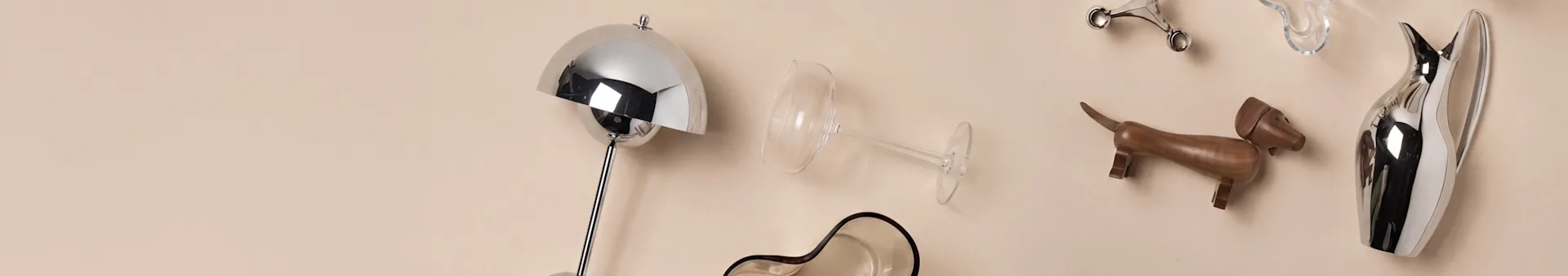 Assortimento di oggetti decorativi per la casa, tra cui una lampada argentata, un bicchiere da vino, un cane in legno e una brocca argentata su sfondo beige.