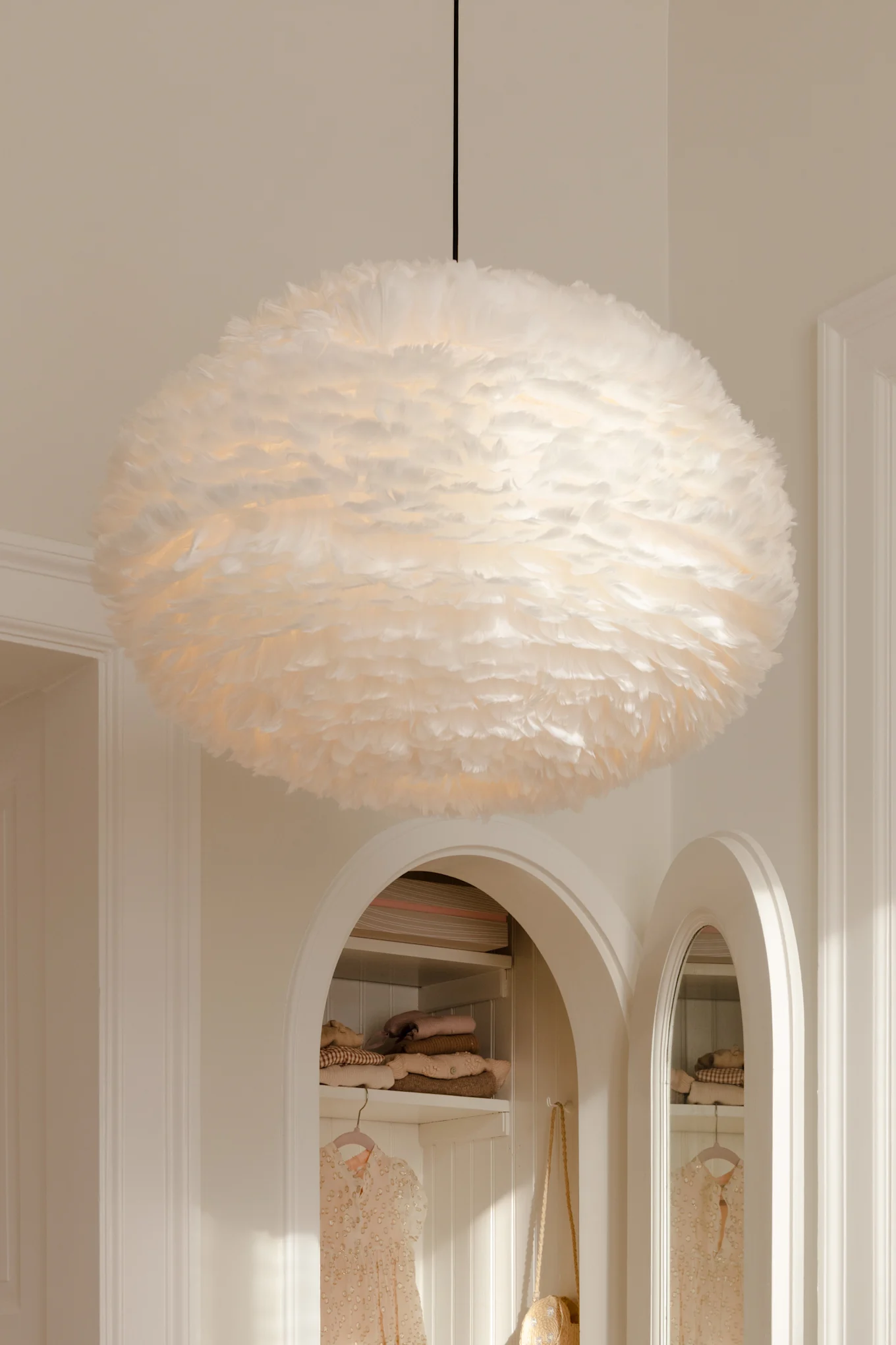 Una grande lampada a sospensione bianca e piumosa pende dal soffitto sopra alcove ad arco con vestiti.