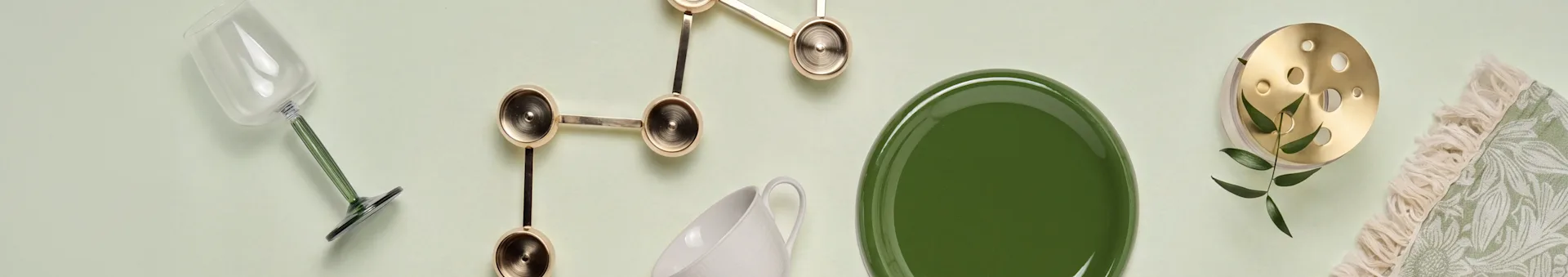 Bicchiere con stelo verde, decoro dorato, tazza bianca, piatto verde, oggetto in ottone con foglie, tessuto verde frangiato.