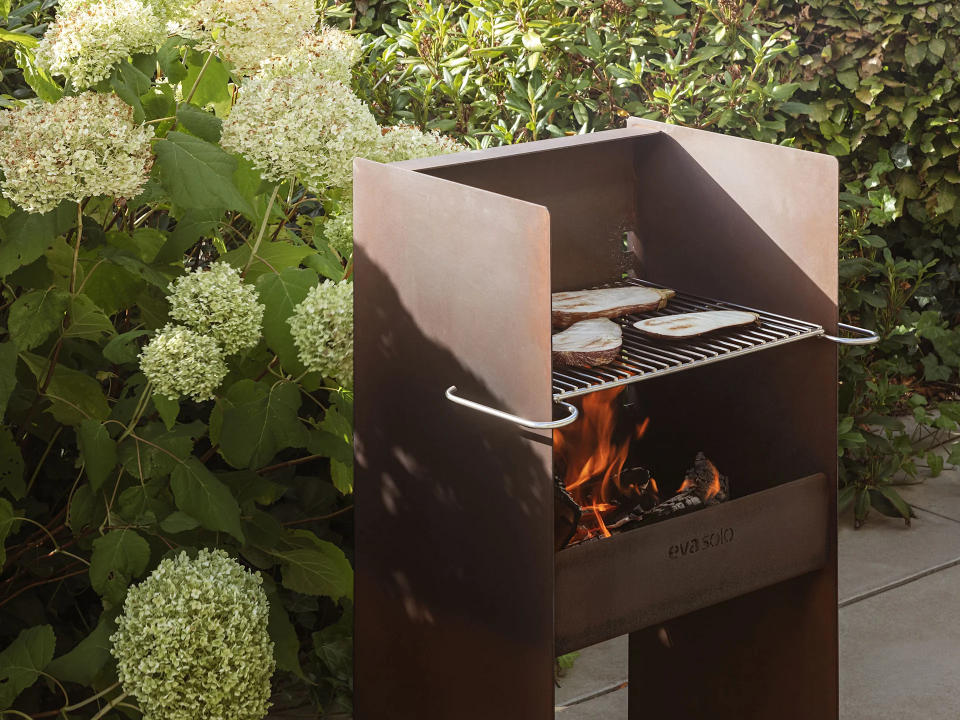 Un barbecue da esterno rustico in metallo con fiamme e fette di melanzane che cuociono sulla griglia.