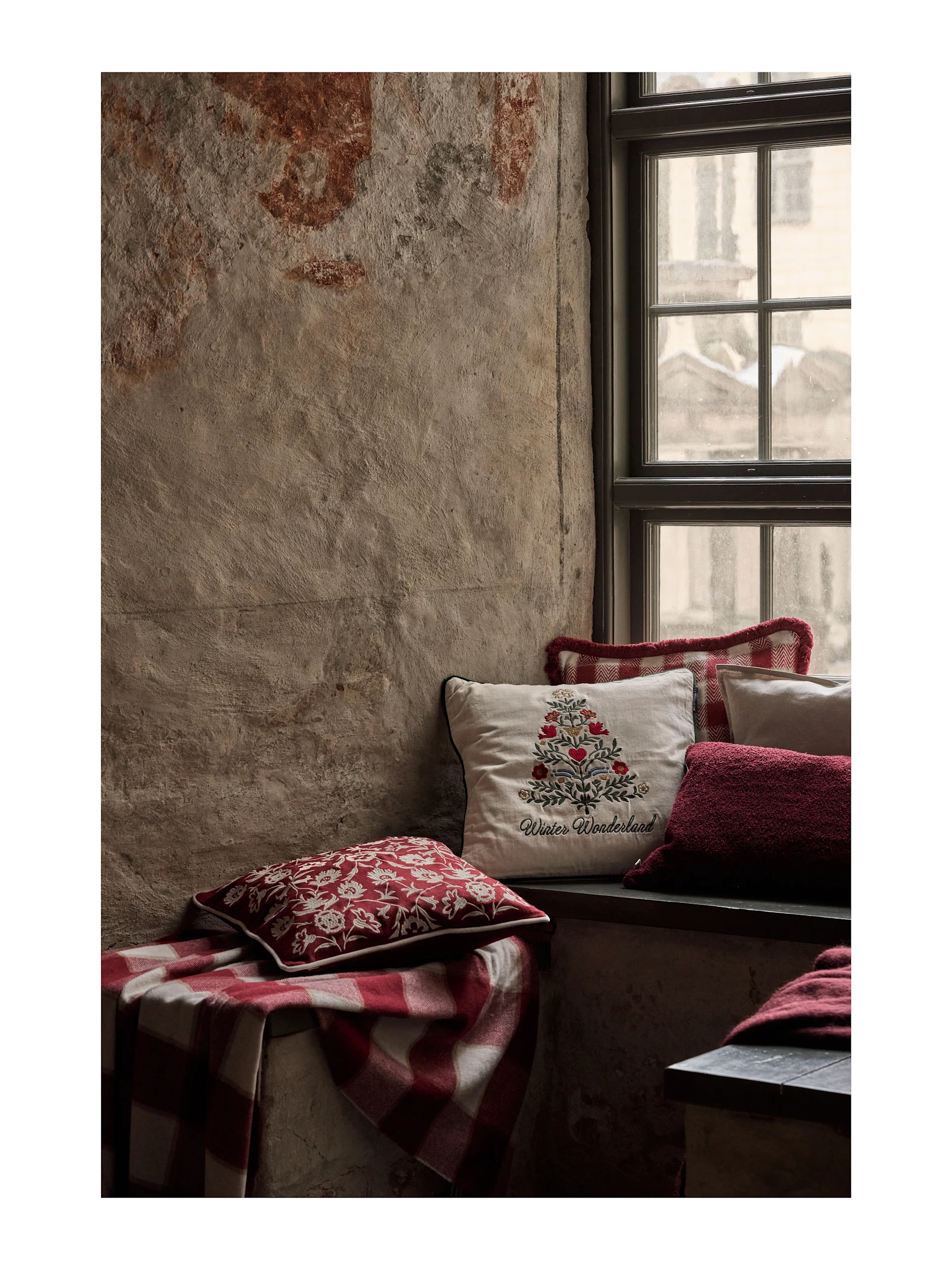 Accogliente sedile a finestra con cuscini festivi rossi e bianchi e una coperta a quadri. Un cuscino color crema presenta il ricamo 'Winter Wonderland'.