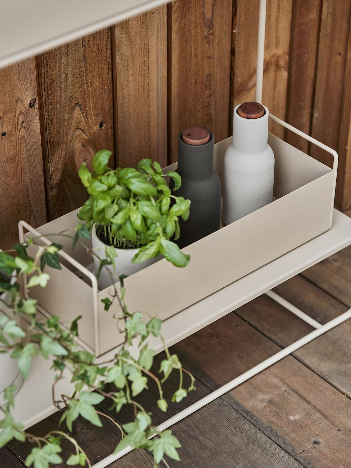 Uno scaffale in metallo beige chiaro con un contenitore che ospita una pianta di basilico, un macinapepe nero e uno bianco su sfondo di legno.