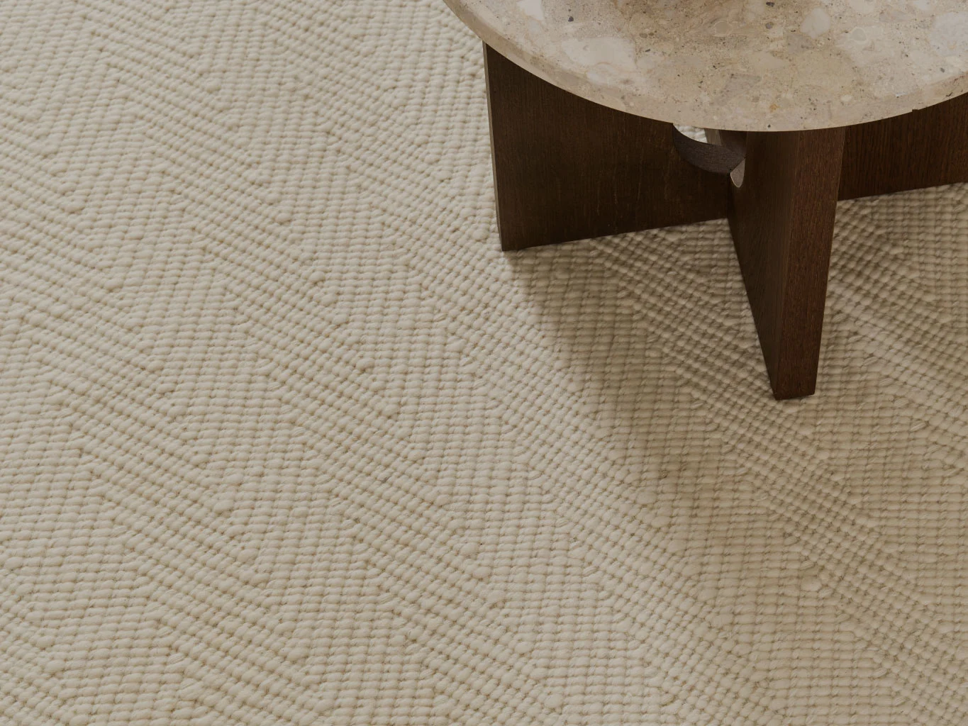 Primo piano di un tappeto color crema testurizzato con motivo a spina di pesce, accanto a un tavolo con piano in pietra.
