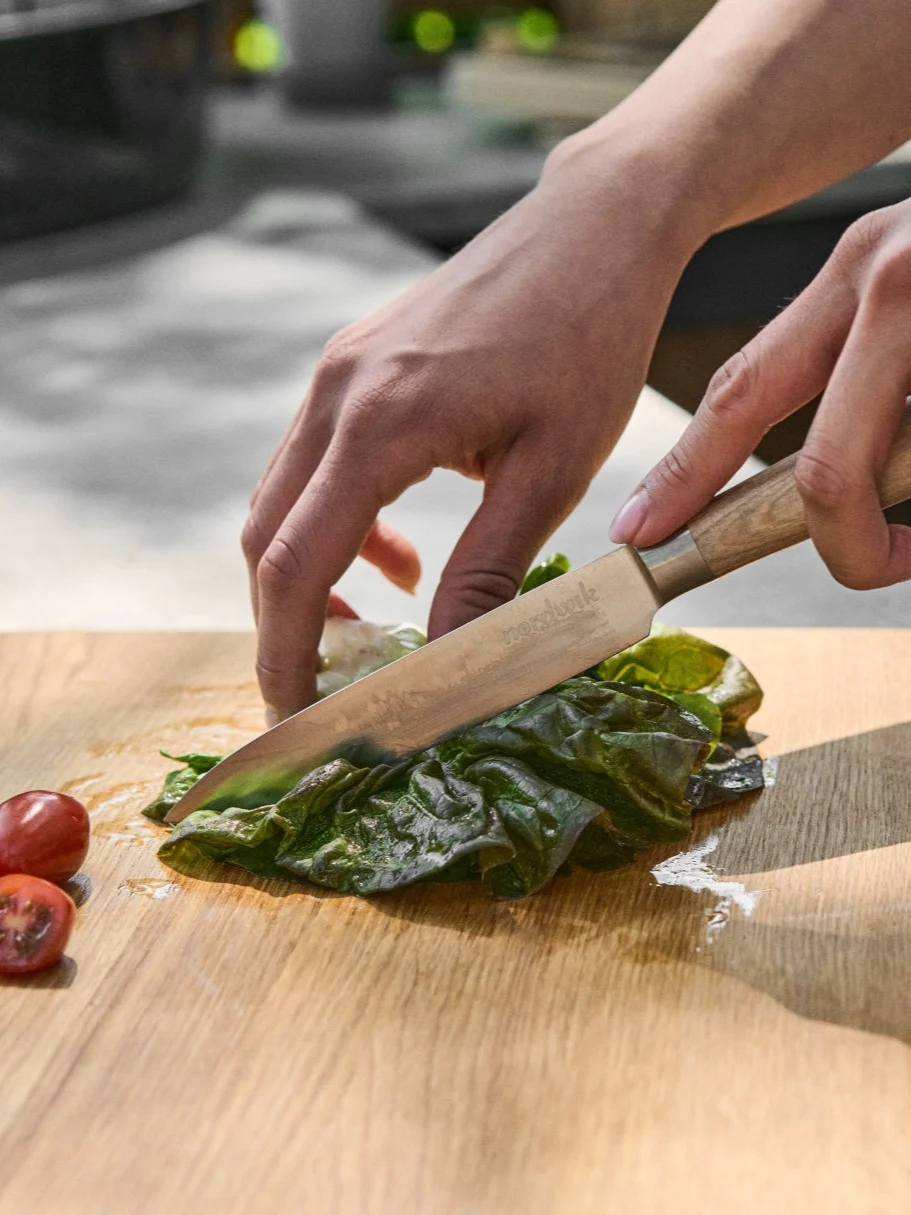 Mani che tagliano lattuga verde fresca su un tagliere di legno con un coltello, accanto a pomodorini tagliati.