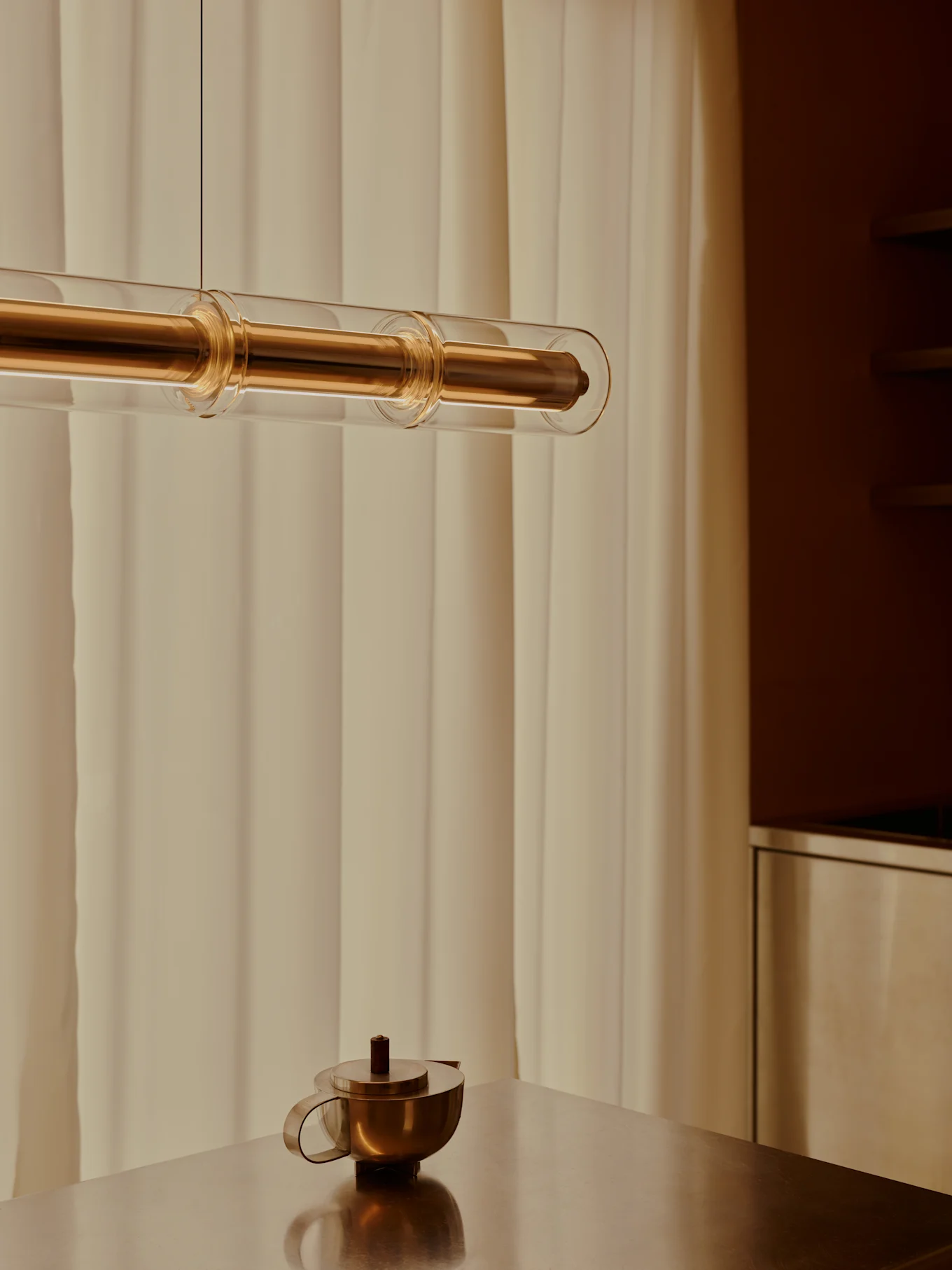 Una moderna lampada a sospensione in ottone e tubi di vetro pende sopra una teiera in ottone su un tavolo metallico, con tende chiare sullo sfondo.