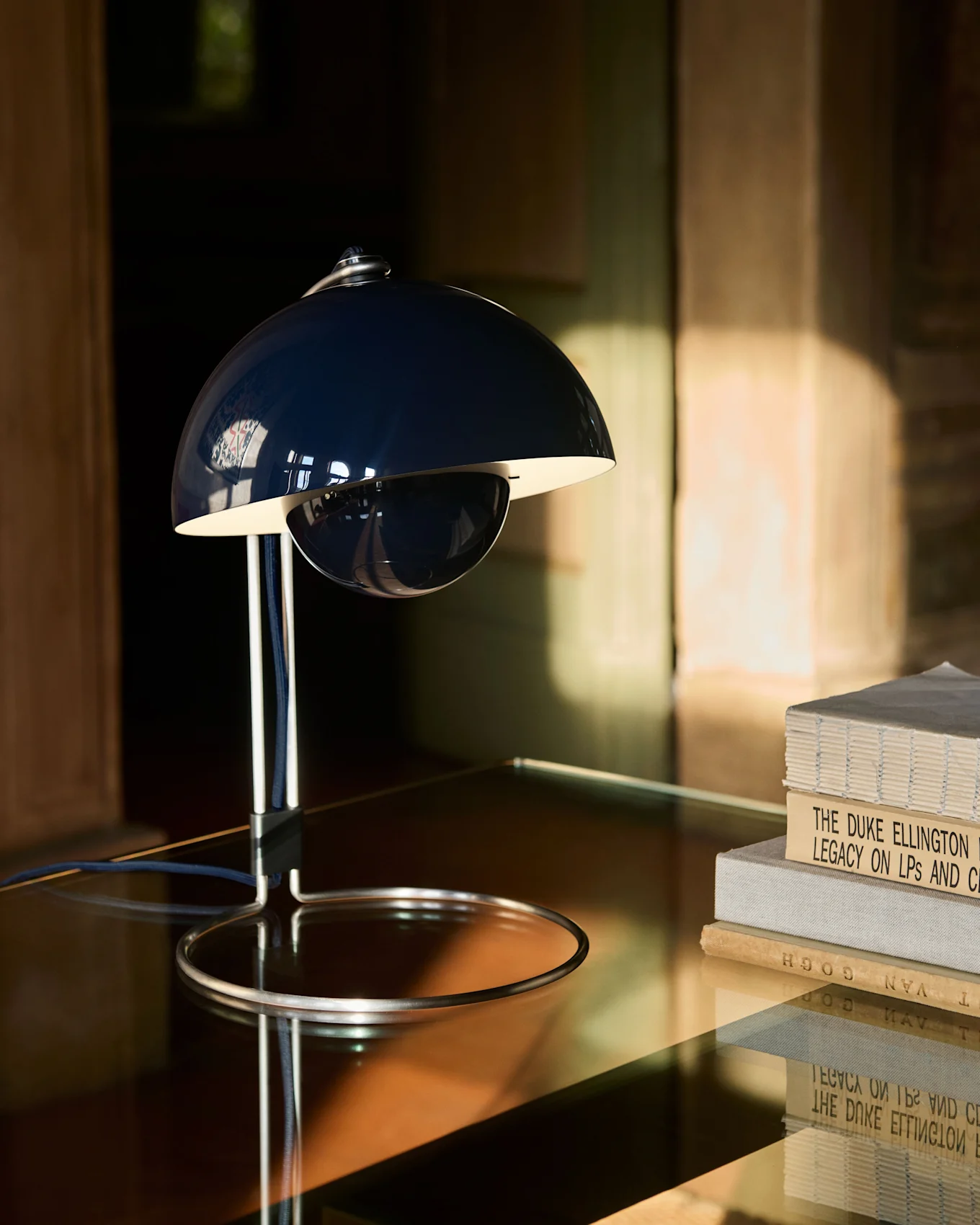 Una lampada da tavolo blu scuro a forma di campana con base cromata si trova su un tavolo riflettente accanto a una pila di libri.