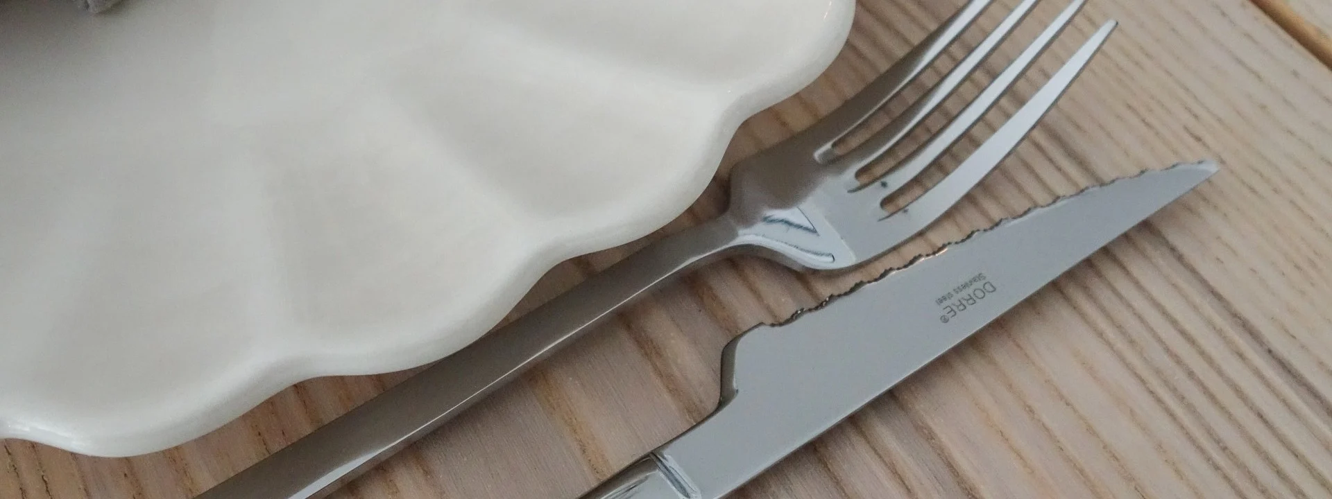 Elegante apparecchiatura: piatto bianco a balze, tovagliolo lino grigio, anello portatovagliolo dorato, forchetta/coltello argento, calice su tavolo legno.