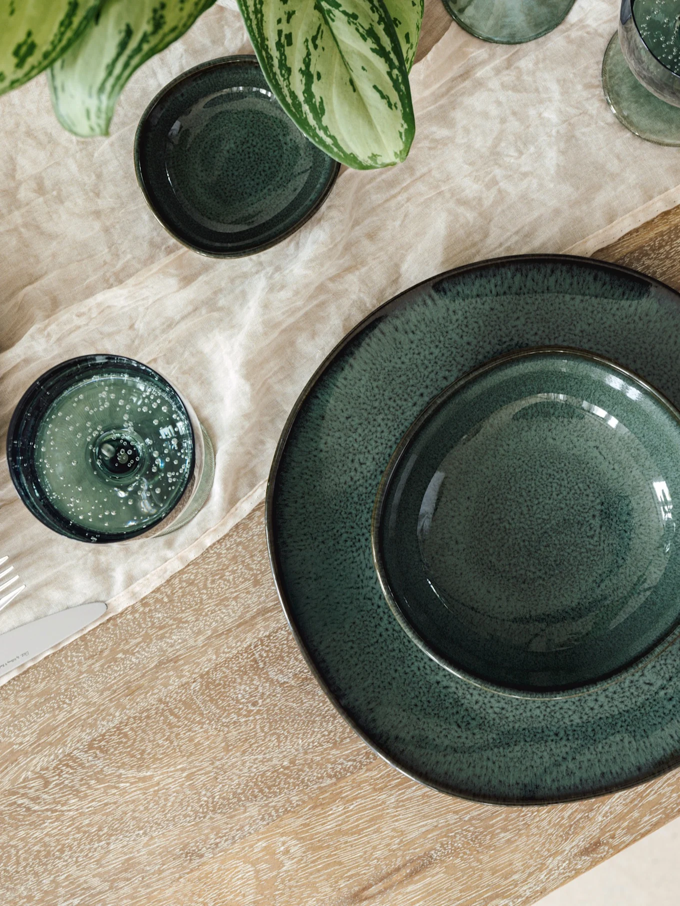 Tavola apparecchiata con piatti, ciotole e bicchieri in ceramica verde scuro su tovaglietta di lino chiaro e tavolo in legno, con pianta.