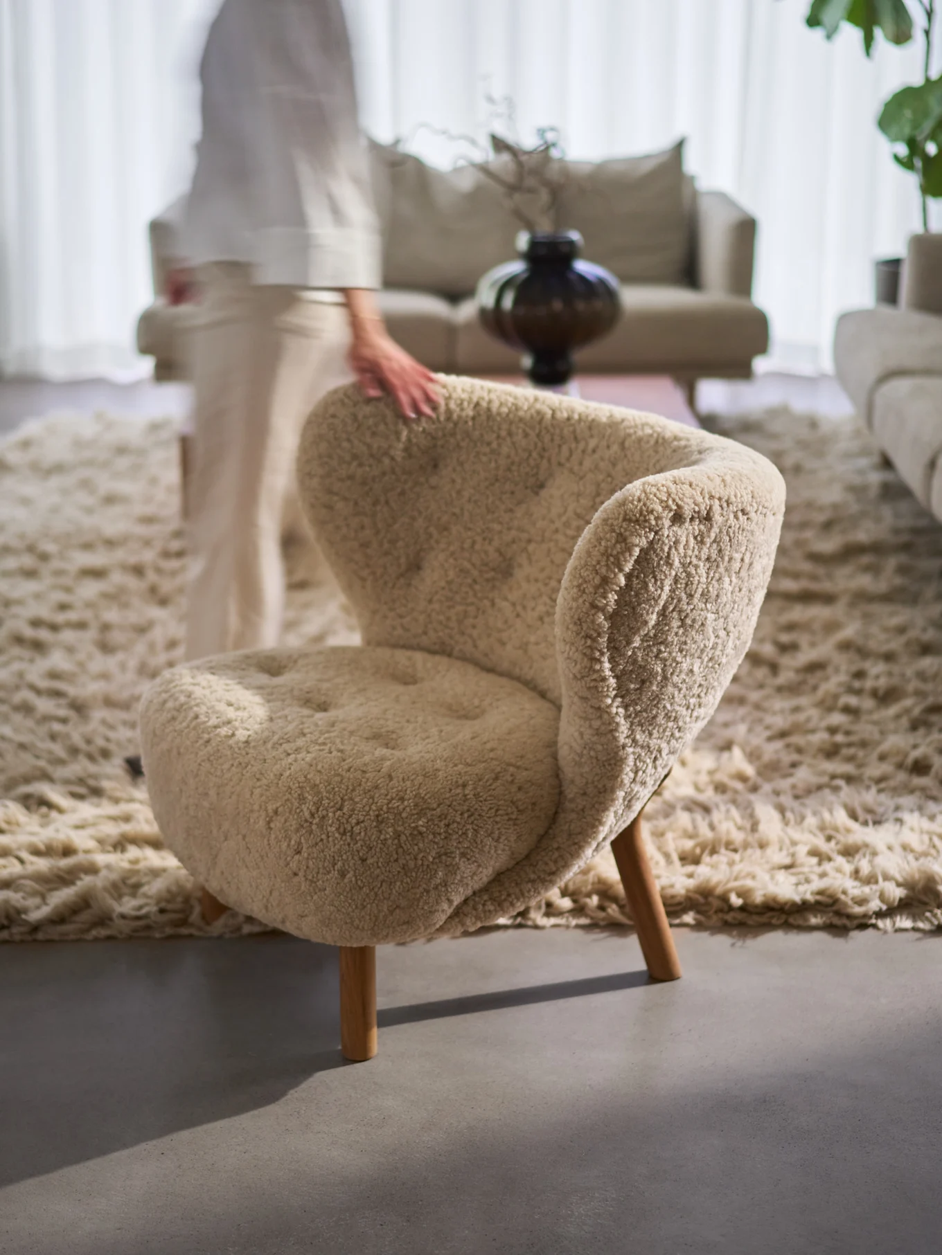La mano di una persona appoggia su una comoda poltrona beige testurizzata con gambe in legno, su un tappeto shaggy in un soggiorno.