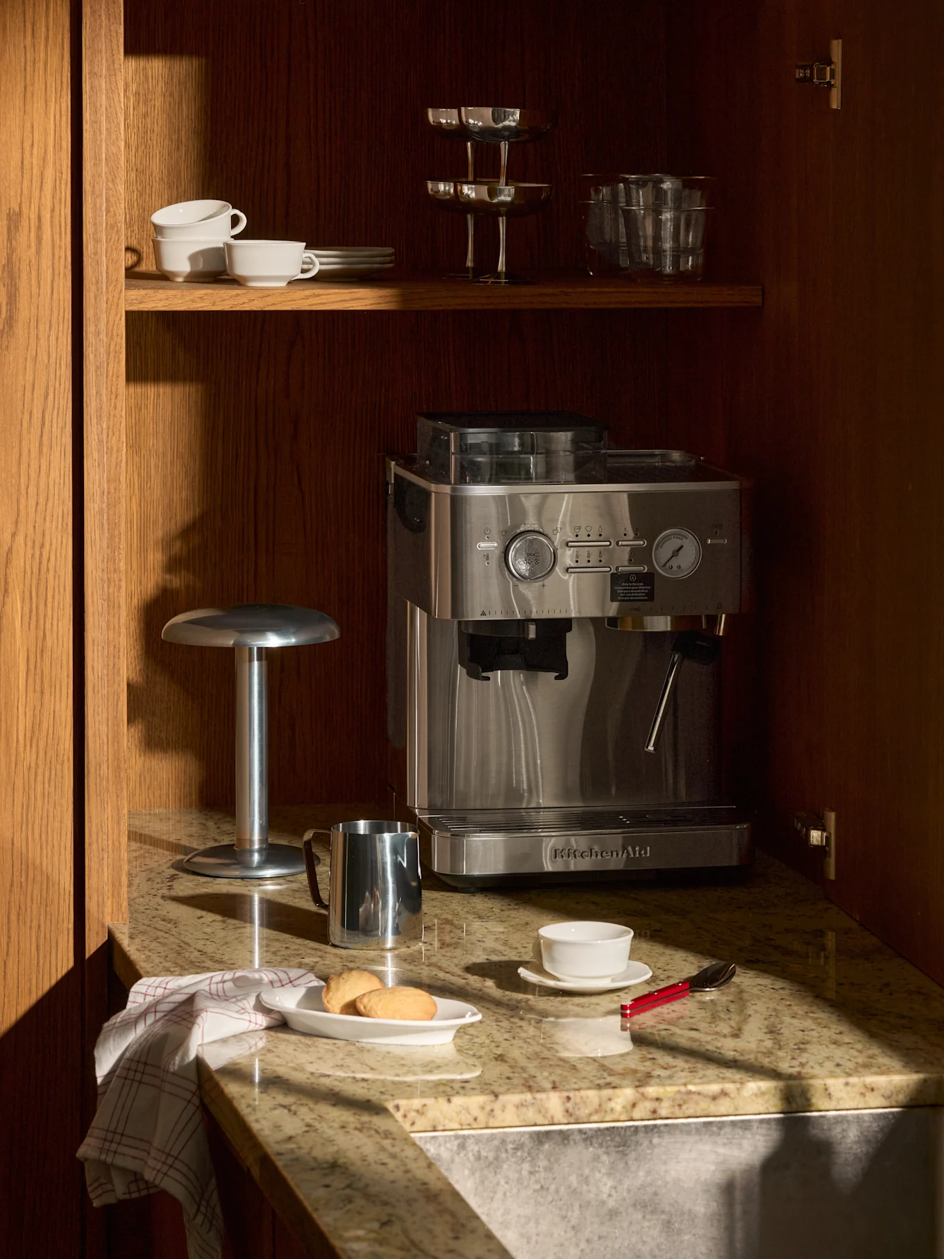 Macchina espresso inox su piano granito in mobile legno. Sopra, tazze, bicchieri. Sul piano, biscotti, tazza, brocca latte.