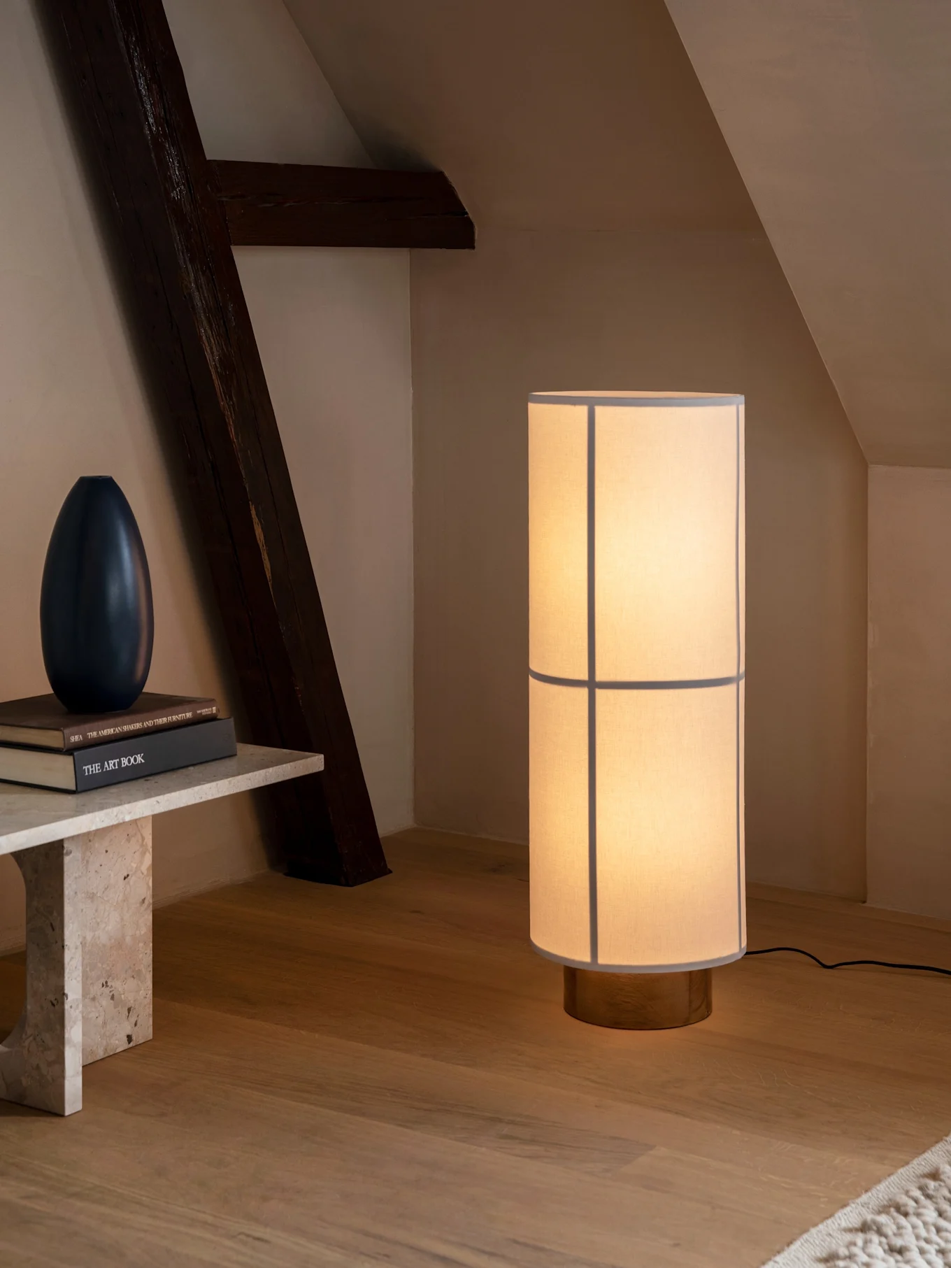 Lampada da terra cilindrica, paralume in tessuto chiaro e base in legno, illumina stanza con panca e trave scura.