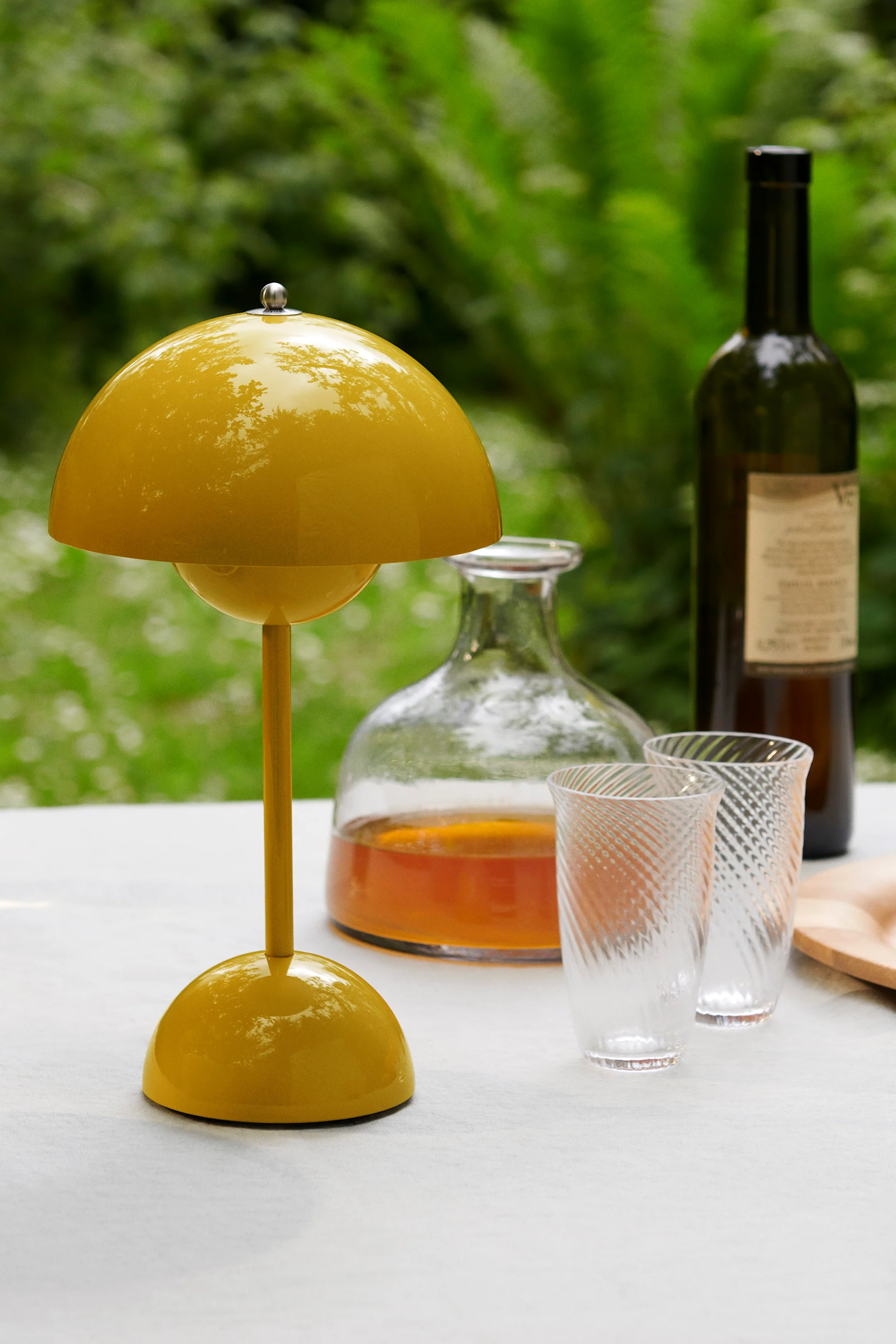 Una lampada da tavolo gialla brillante a forma di fungo su un tavolo bianco, con una caraffa, due bicchieri modellati e una bottiglia di vino dietro, su sfondo verde.