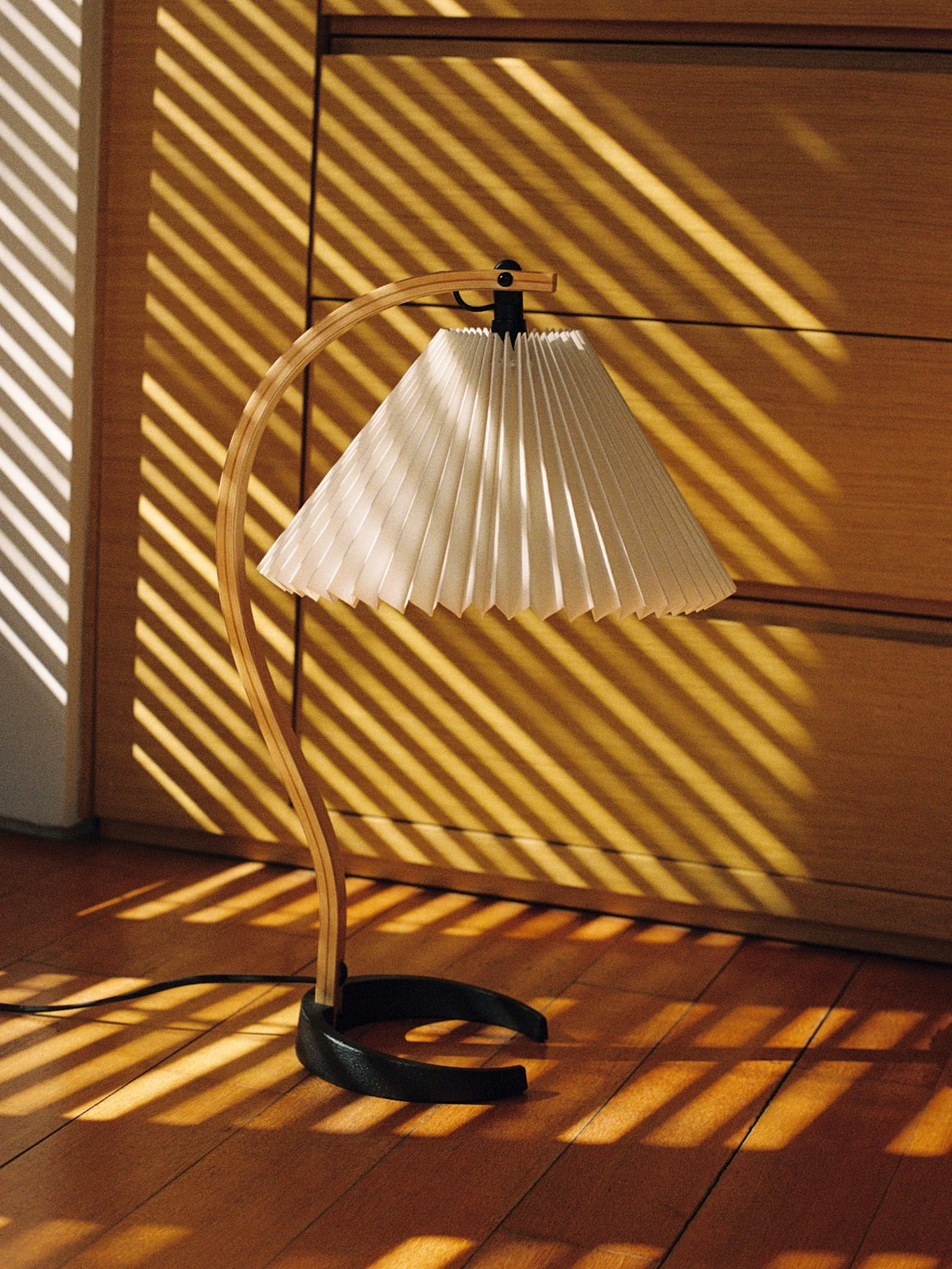 Lampada da tavolo con base in legno curvato e paralume plissettato, illuminata da strisce di luce diagonali su pavimento in legno.
