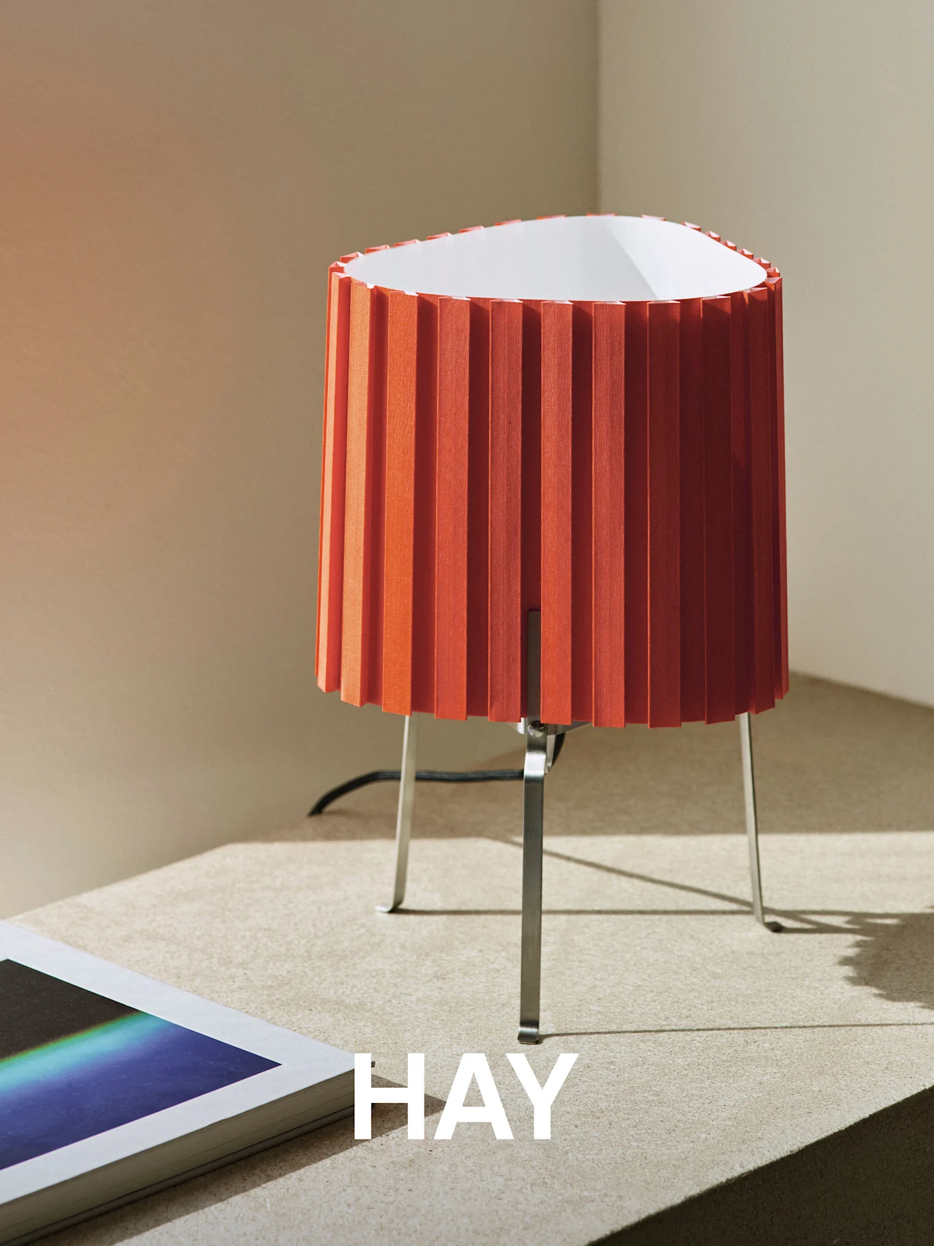 Lampada da tavolo plissettata arancione con base in metallo su una superficie ruvida, accanto a un libro. La parola 'HAY' è sovrapposta.