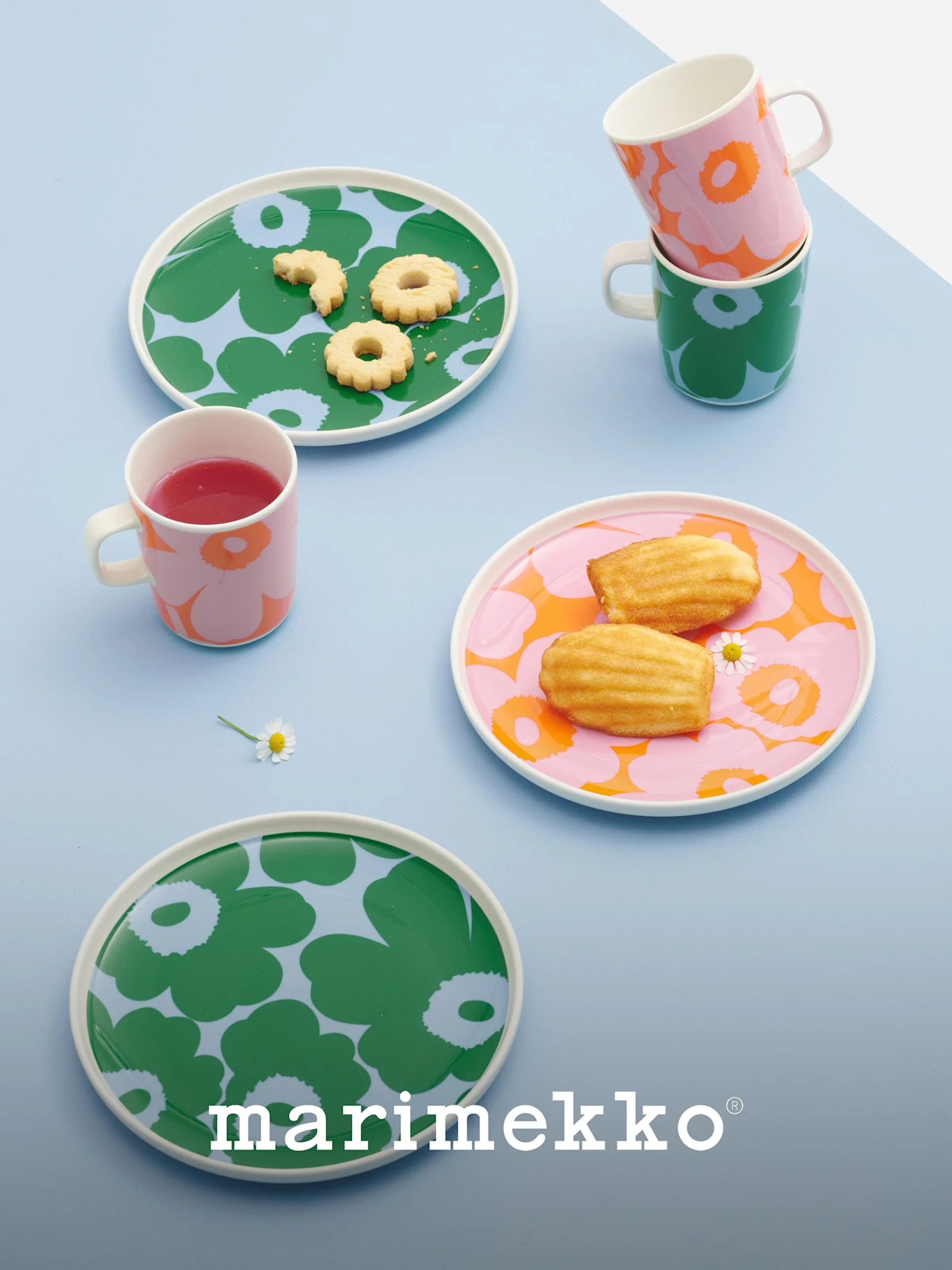 Piatti e tazze Marimekko con audaci motivi floreali in verde, rosa e arancione, contenenti biscotti, madeleine e una bevanda.