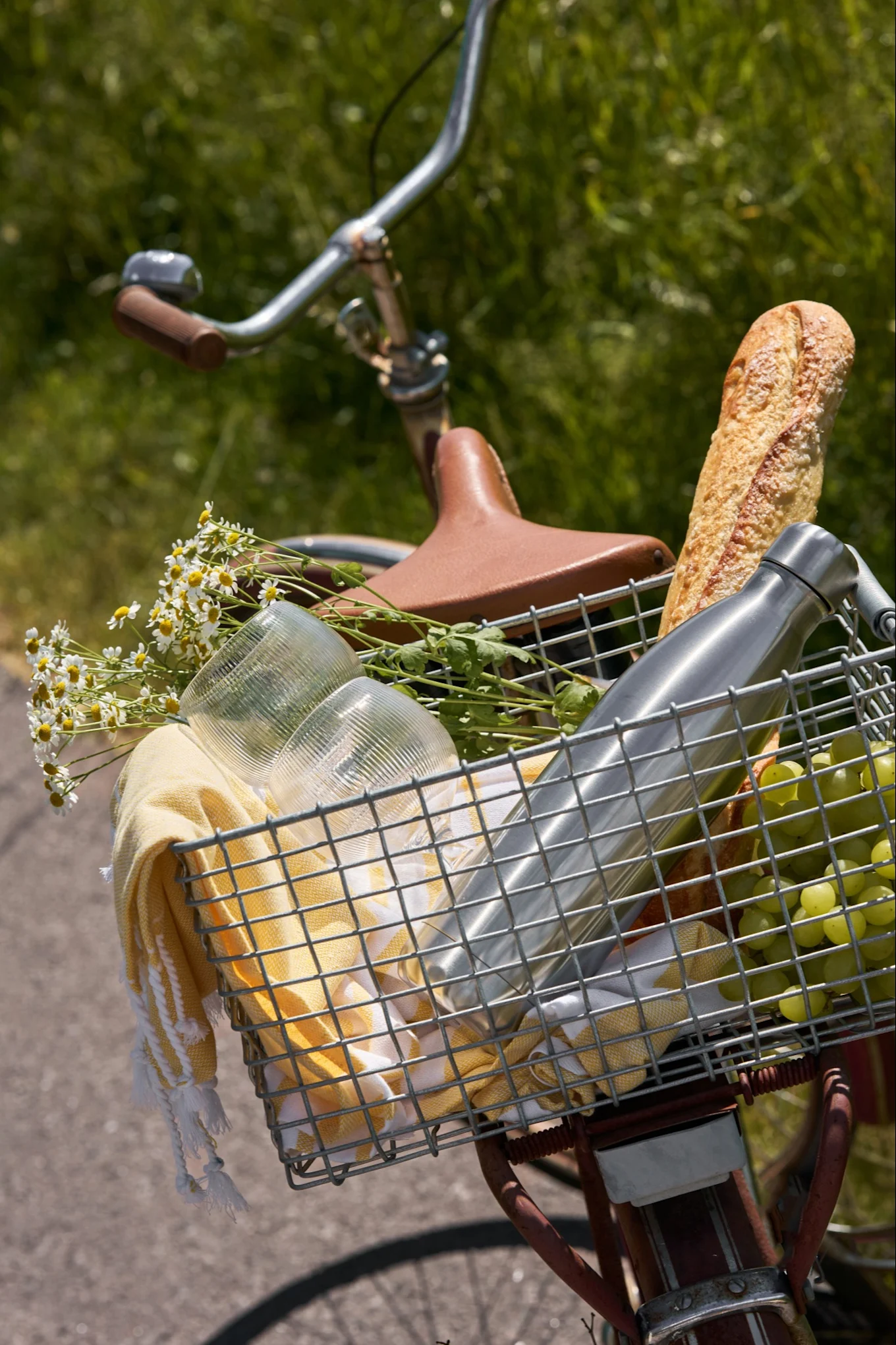 Cesto anteriore di bicicletta con articoli da picnic: baguette, borraccia, uva, bicchieri, asciugamano giallo e margherite bianche.