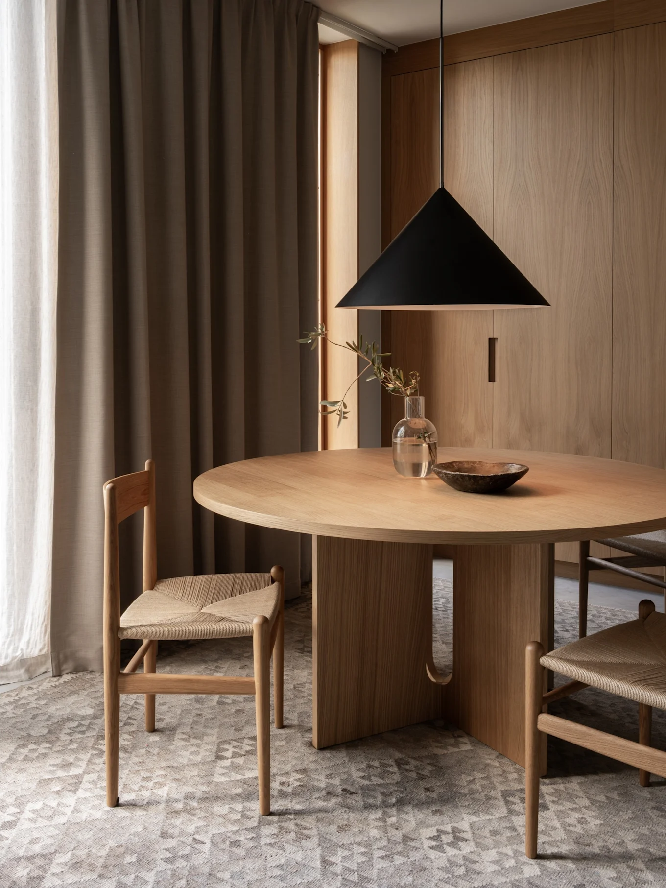 Una zona pranzo minimalista con un tavolo rotondo in legno, due sedie con sedili intrecciati, una lampada a sospensione nera e pareti in legno.