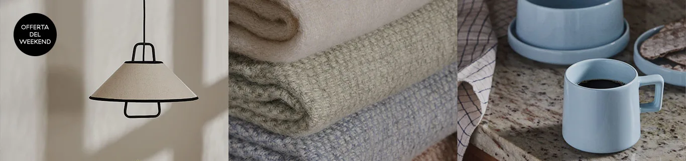 Offerta del weekend: lampada a sospensione beige, pile di coperte pastello e tazza da caffè blu su bancone.
