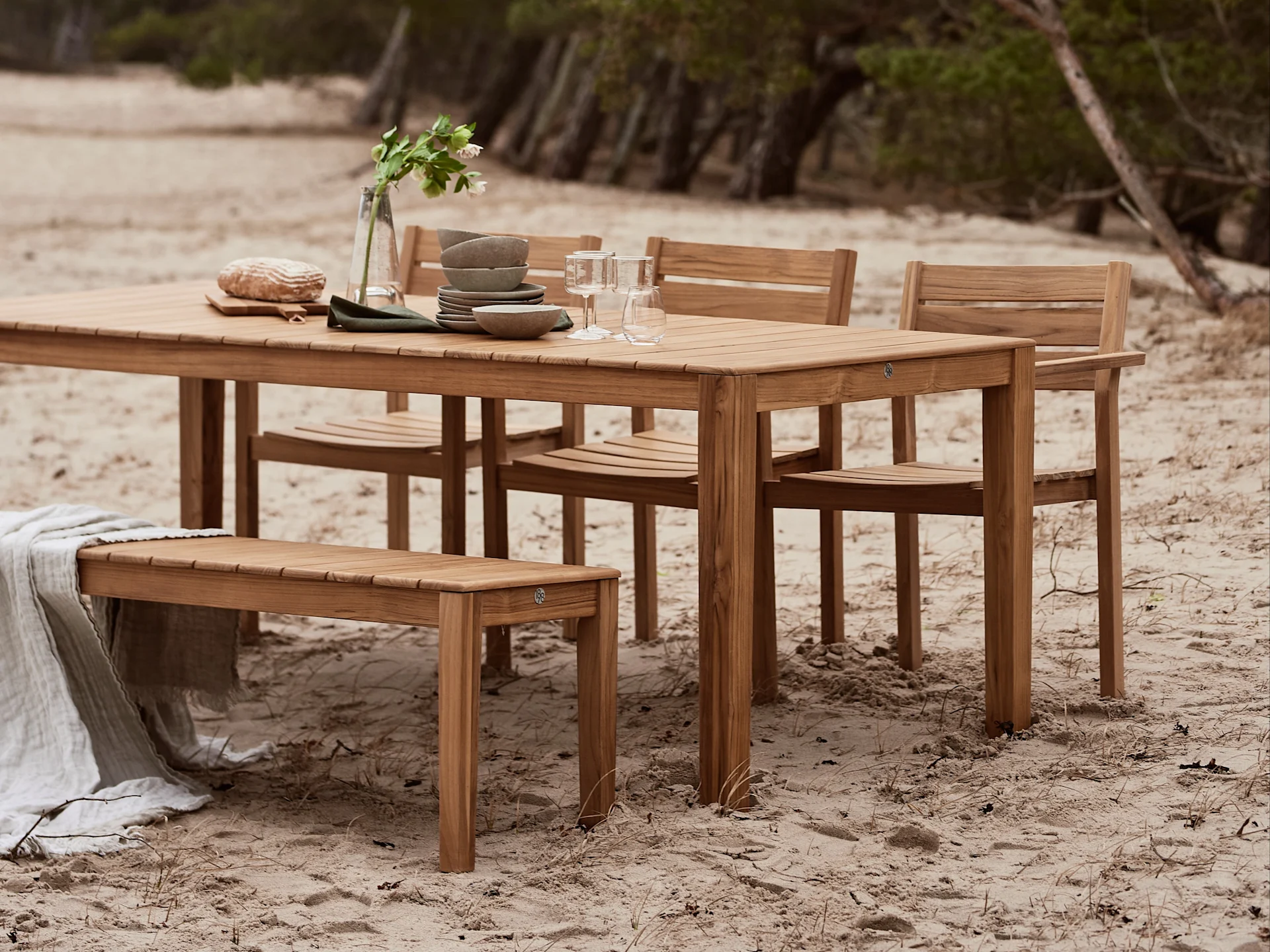 Set da pranzo esterno in legno con tavolo, sedie e panca, allestito con stoviglie e una coperta su una spiaggia sabbiosa.