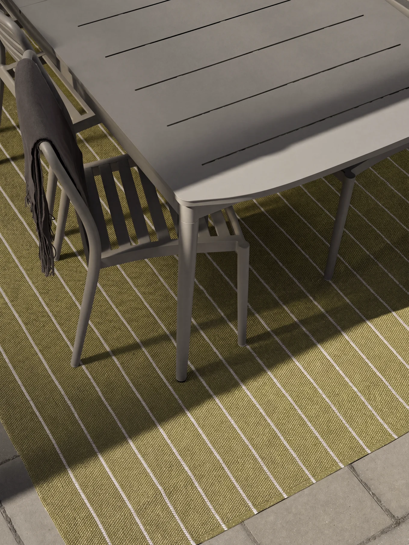 Tavolo e sedie da pranzo grigi per esterni su un tappeto a righe verdi e bianche. Una coperta grigio scuro è drappeggiata su una sedia.