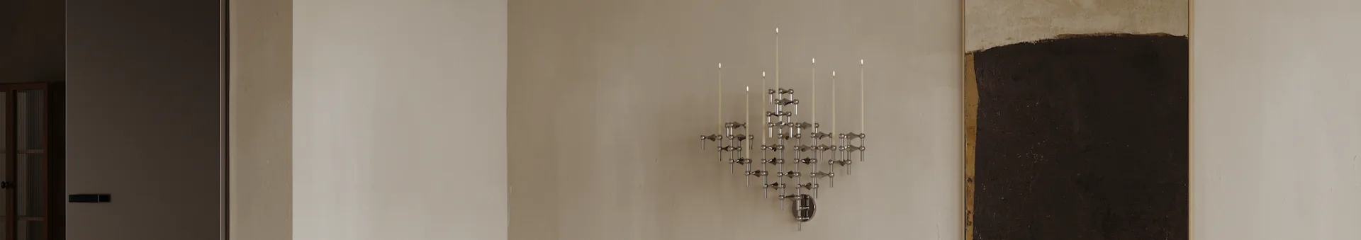 Un candelabro modulare geometrico cromato moderno con diverse candele bianche accese montato su una parete chiara.