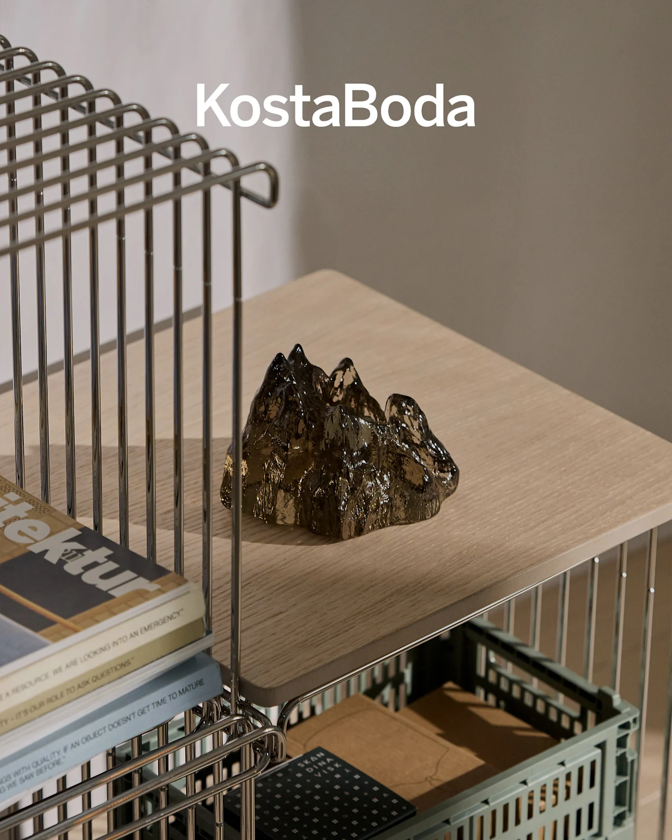Marchio KostaBoda sopra una scultura in vetro scura, trasparente, a forma di montagna su una mensola di legno chiaro.