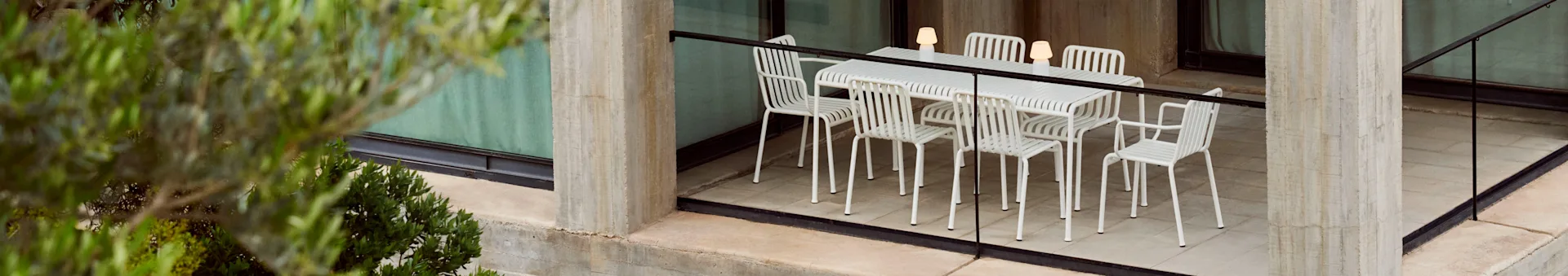 Un set da pranzo all'aperto con un tavolo bianco a doghe e sei sedie abbinate, illuminato da tre lampade portatili, su un patio in cemento con piante.