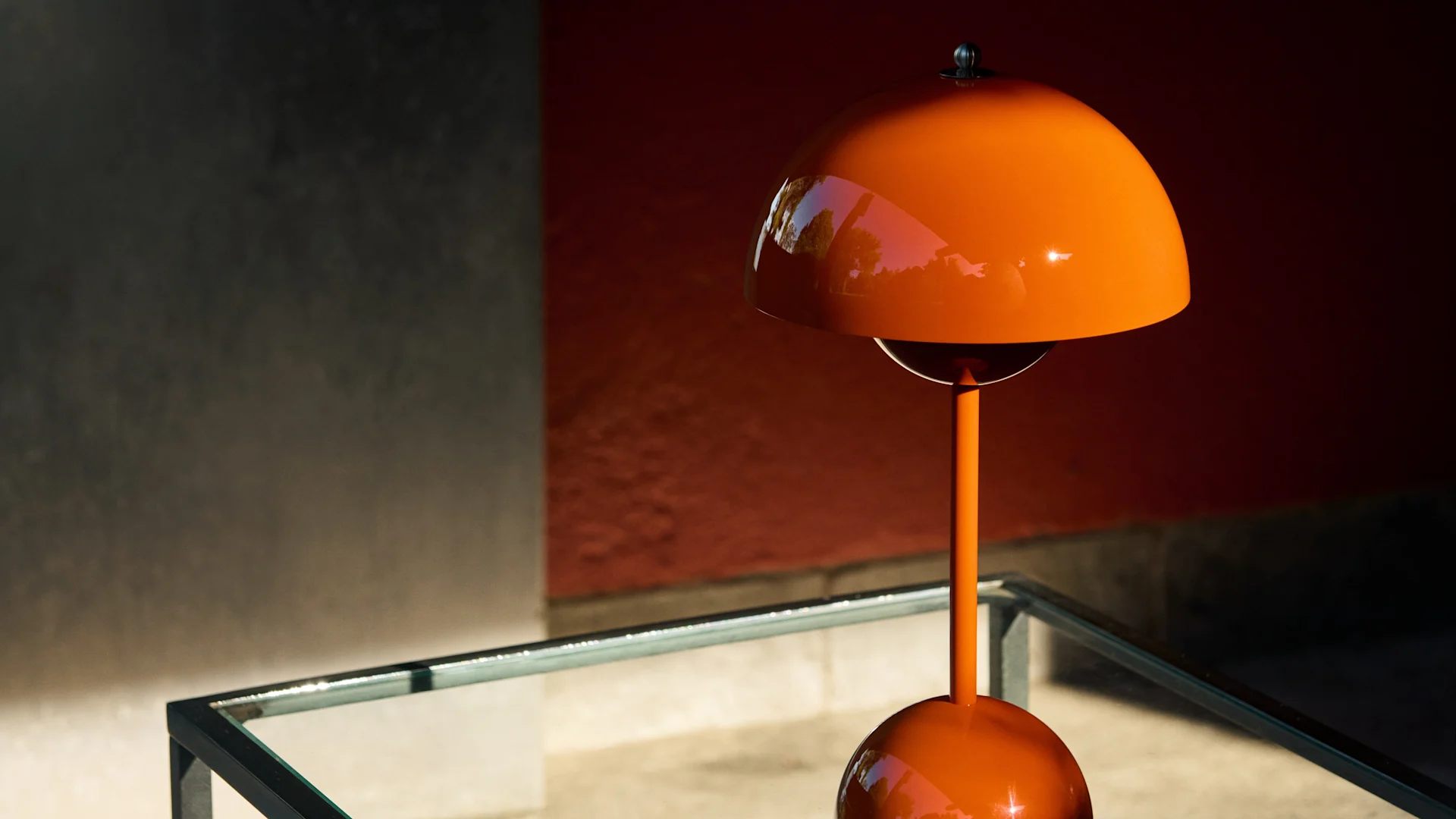 Lampada da tavolo arancione lucida a forma di fungo su un tavolo di vetro contro una parete rossa e grigia, con luce solare a chiazze.