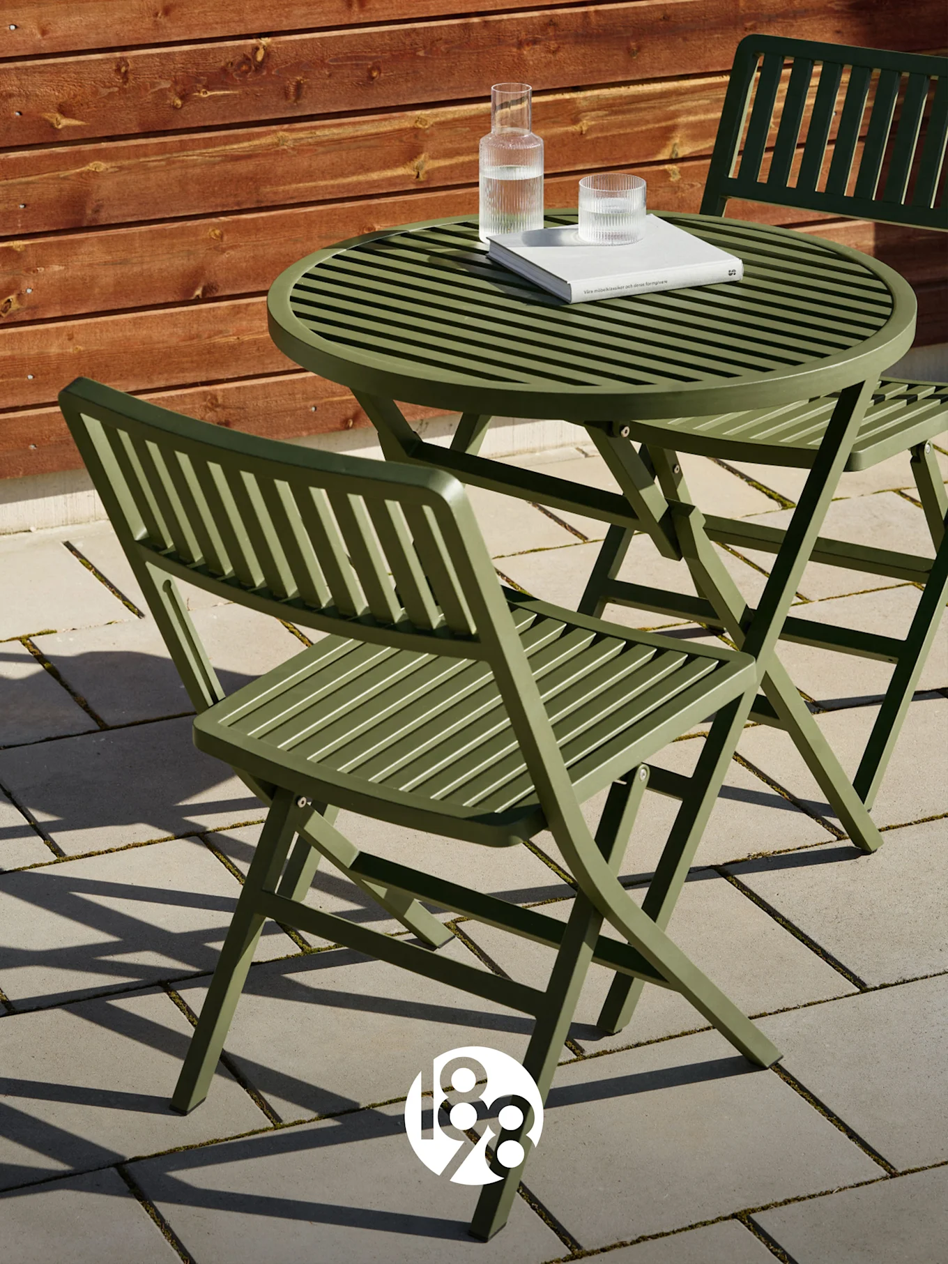 Due sedie pieghevoli verdi a doghe e un tavolo abbinato su un patio con un libro e bicchieri d'acqua.