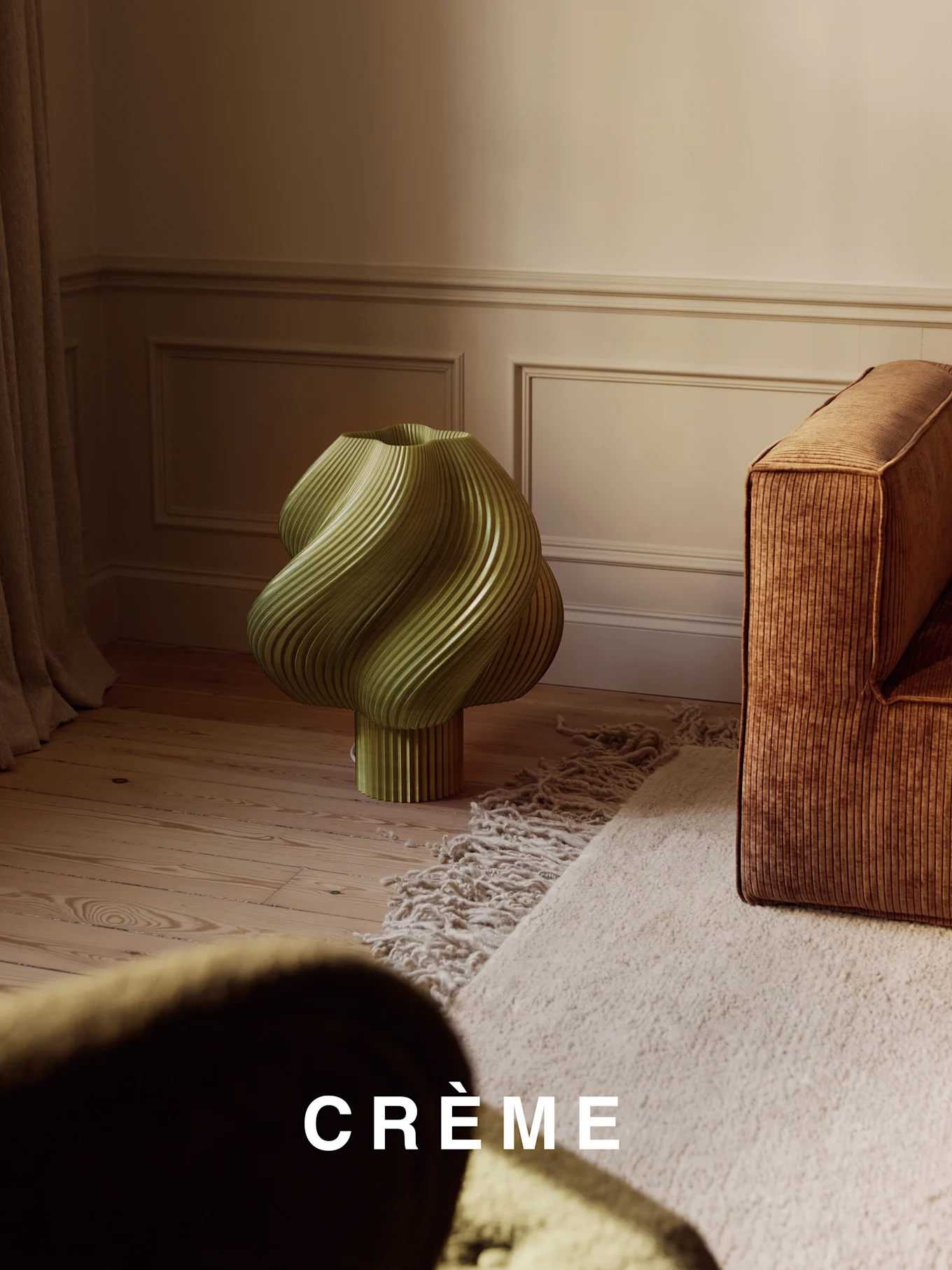 Una lampada verde chiaro, attorcigliata e a coste, su un pavimento in legno, accanto a un divano a coste marrone e un tappeto shaggy beige.