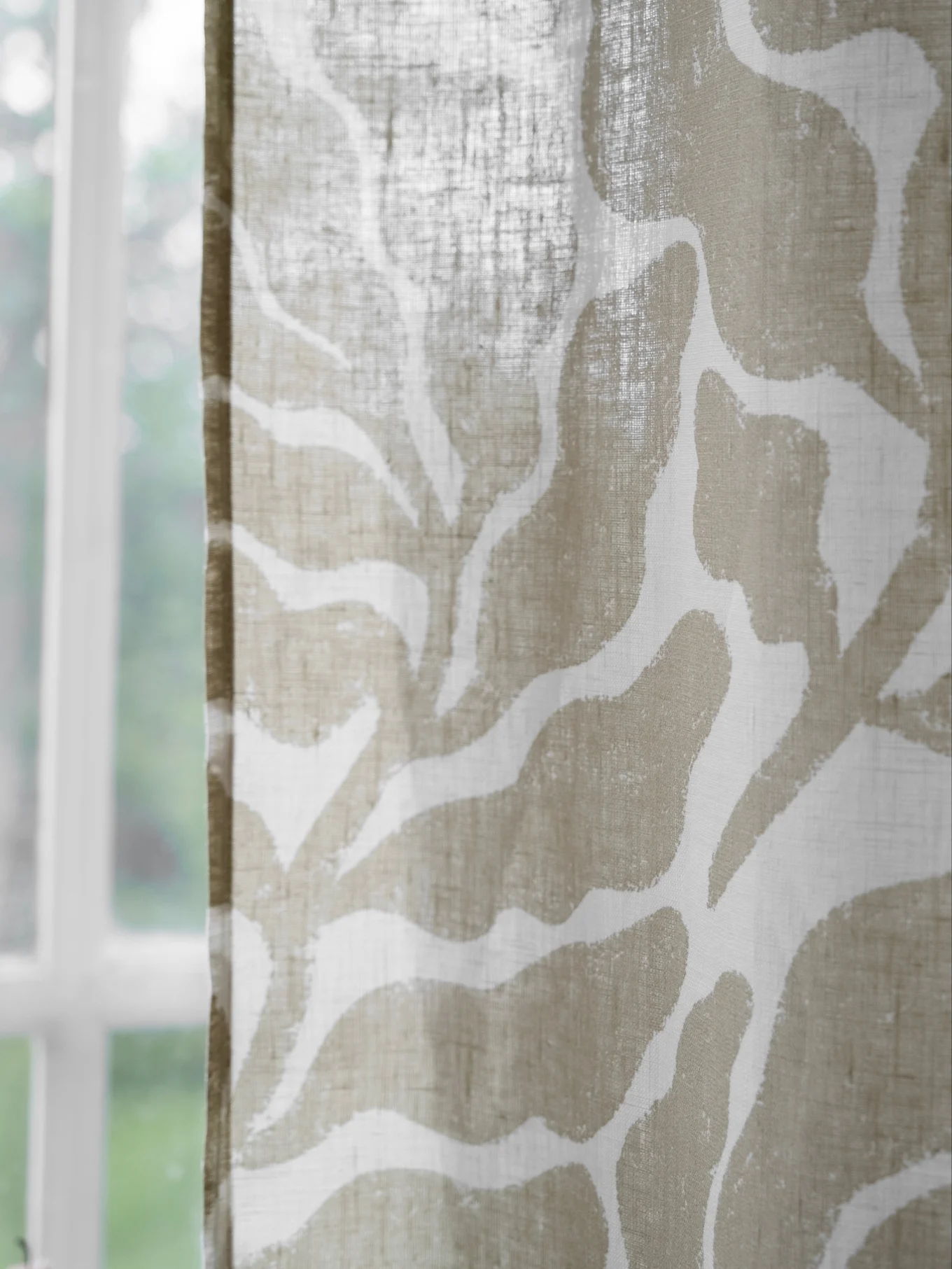 Primo piano di una tenda trasparente con motivo astratto beige, con finestra sfocata e verde sullo sfondo.
