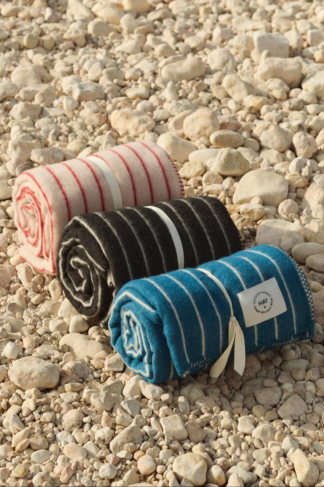 Tre coperte a righe arrotolate in rosa, nero e blu, legate con nastri su una spiaggia di ciottoli.