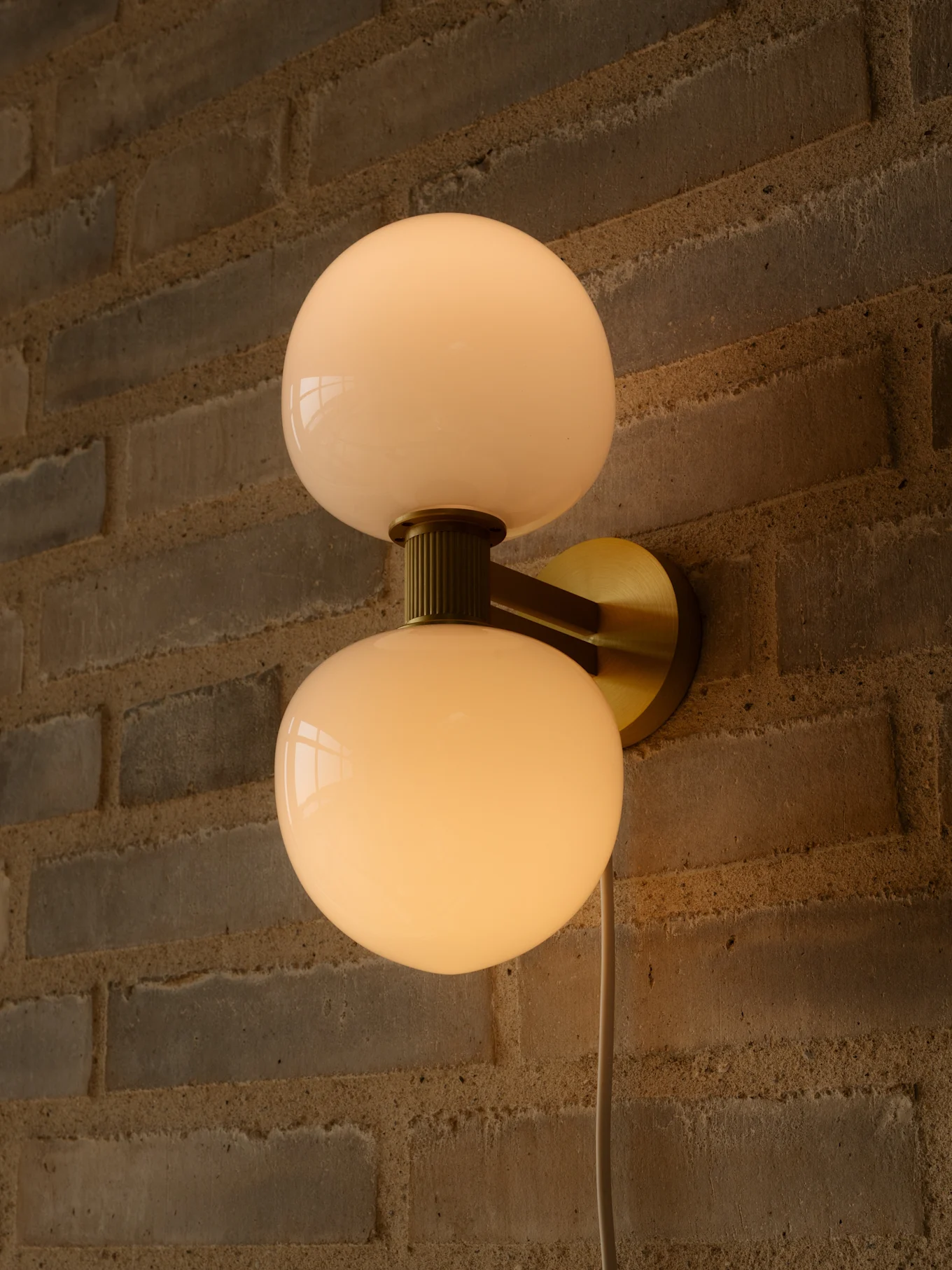 Lampada da parete con due paralumi sferici in vetro bianco latte e montatura in ottone su una parete di mattoni.