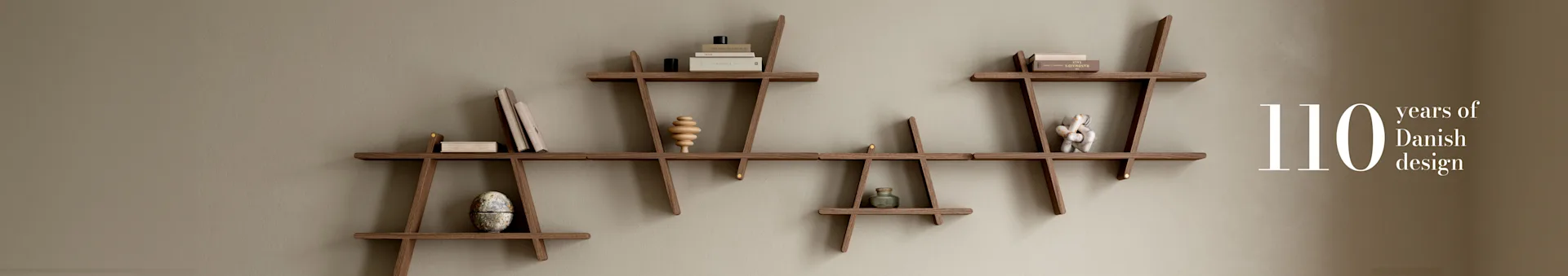 Sistema di mensole modulari in legno su una parete marrone chiaro, con libri e decorazioni. Testo: "110 anni di design danese".