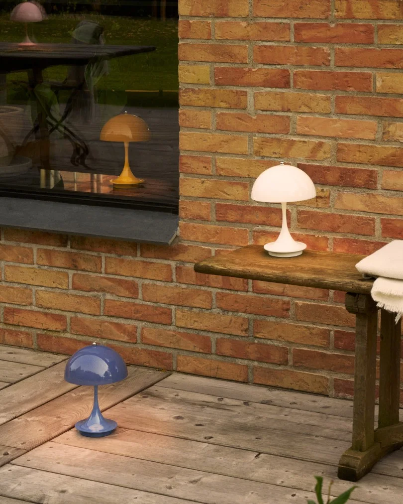 Tre lampade a forma di fungo in bianco, blu e giallo su un patio in legno vicino a un muro di mattoni e una finestra.