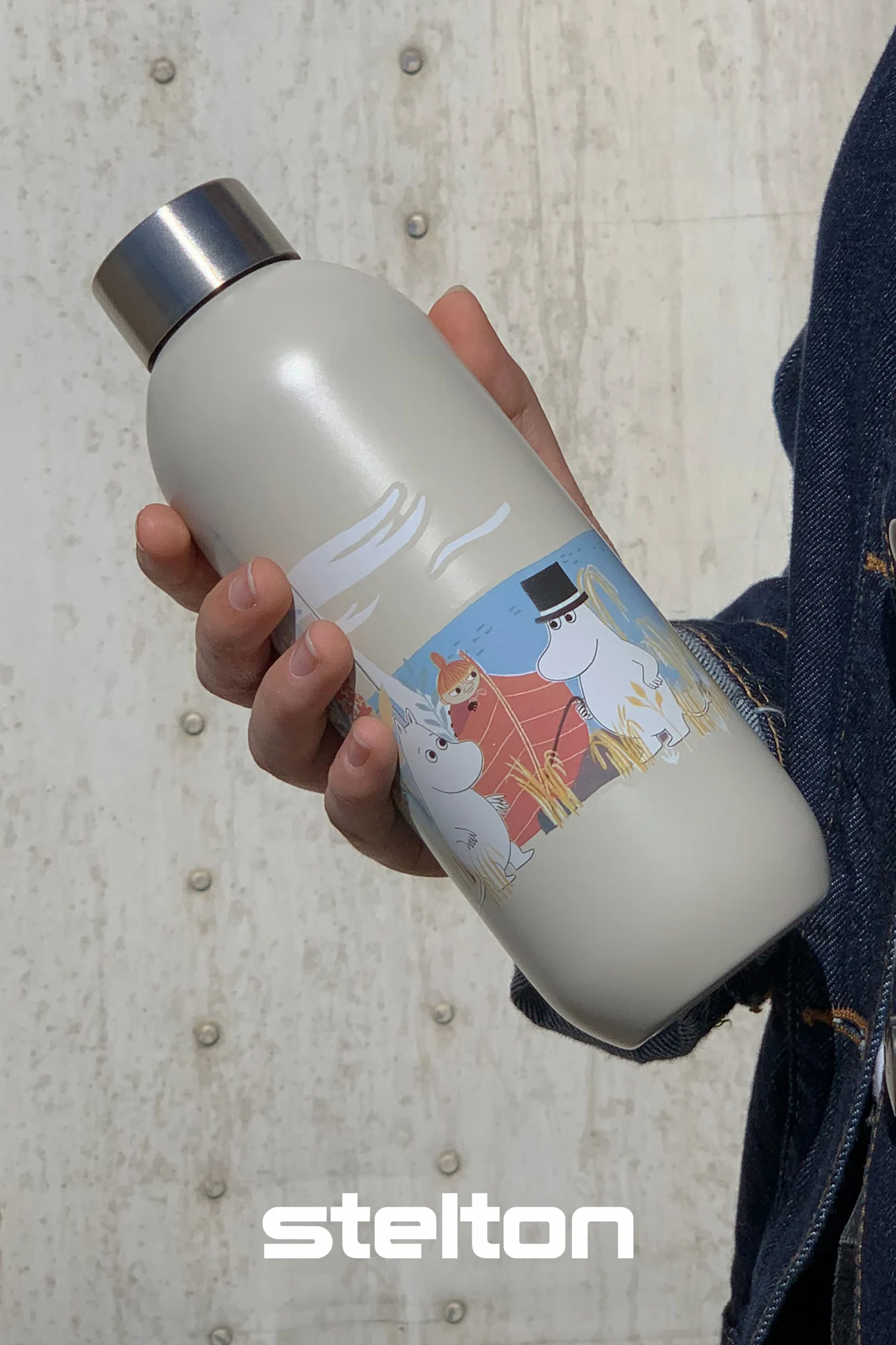 Persona che tiene in mano una bottiglia d'acqua Moomin grigio chiaro con tappo argentato, raffigurante Moomintroll, Piccola My e Moominpappa in barca.