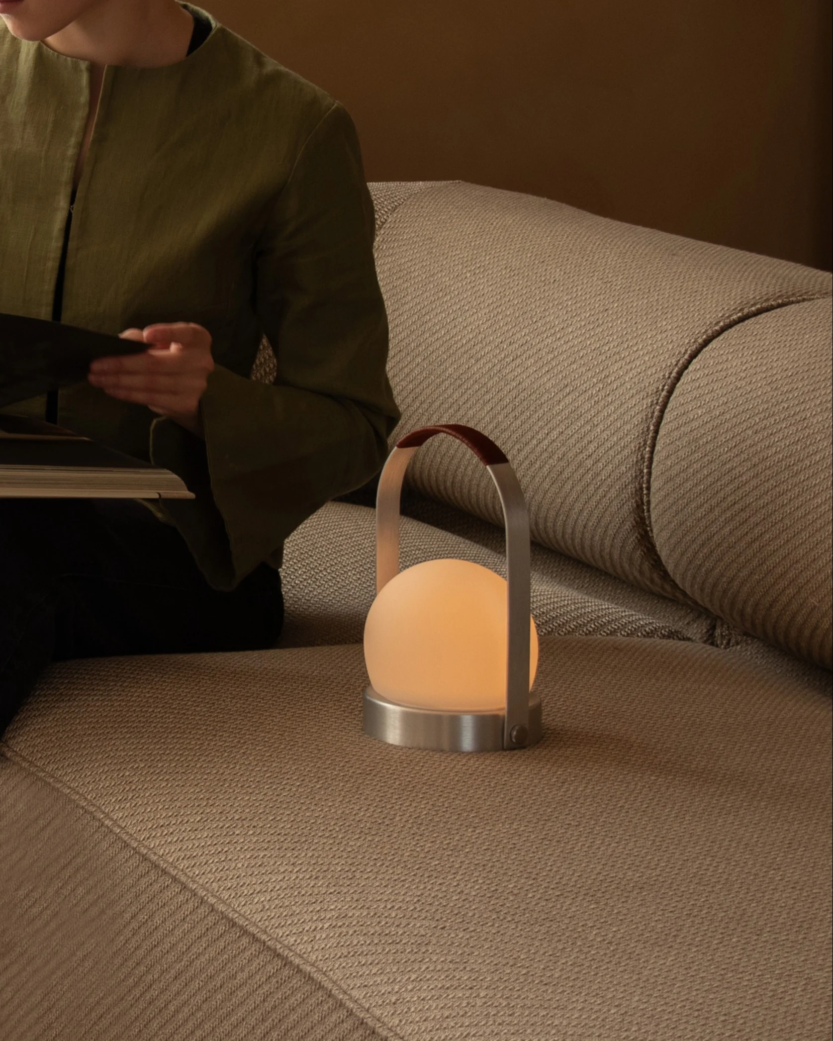 Una persona legge un libro su un divano beige accanto a una lampada portatile sferica luminosa con manico in pelle.