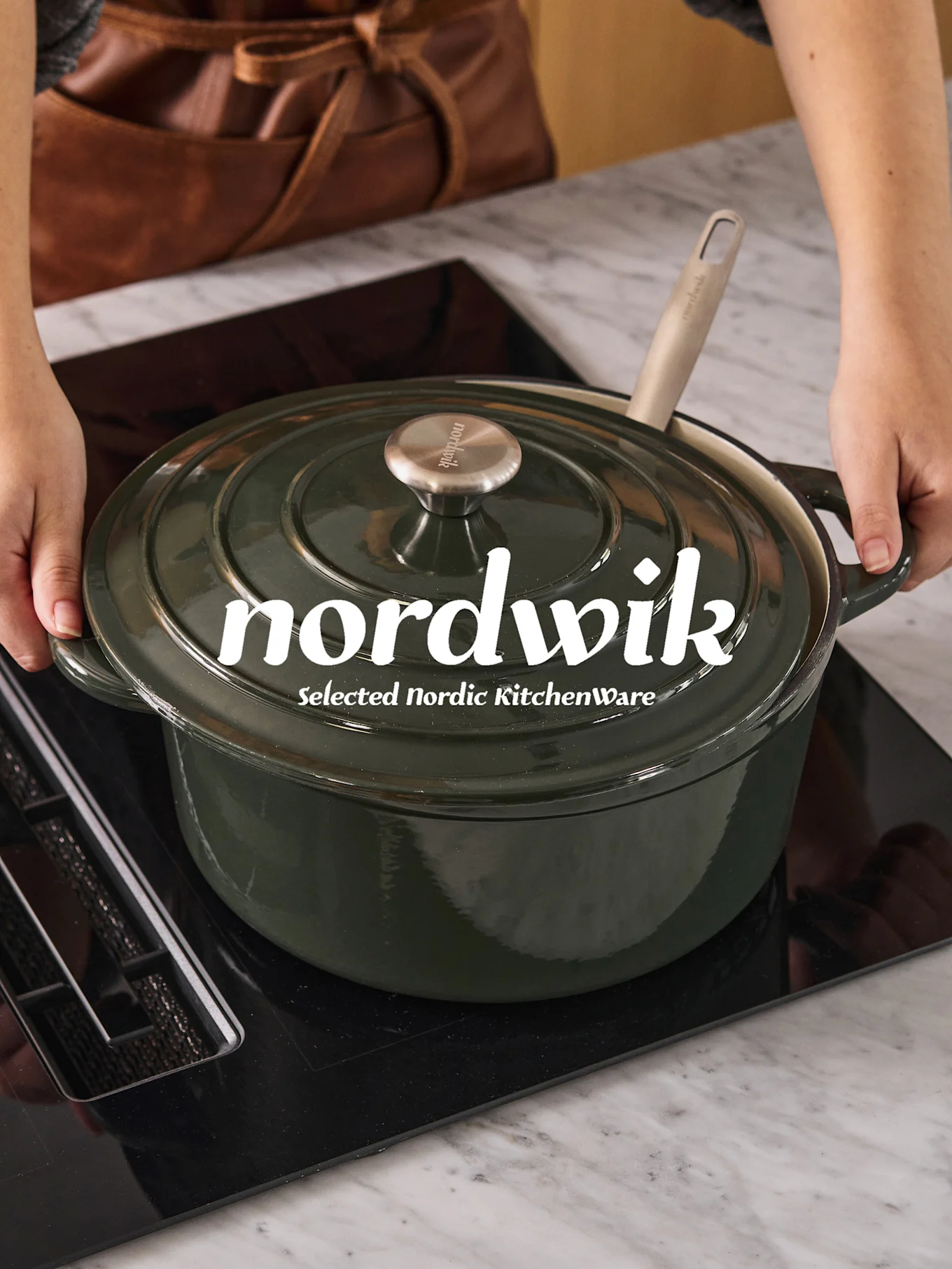 Mani che posizionano una casseruola olandese verde scuro con un cucchiaio chiaro su un piano cottura a induzione nero. 'nordwik' è visibile sul coperchio.
